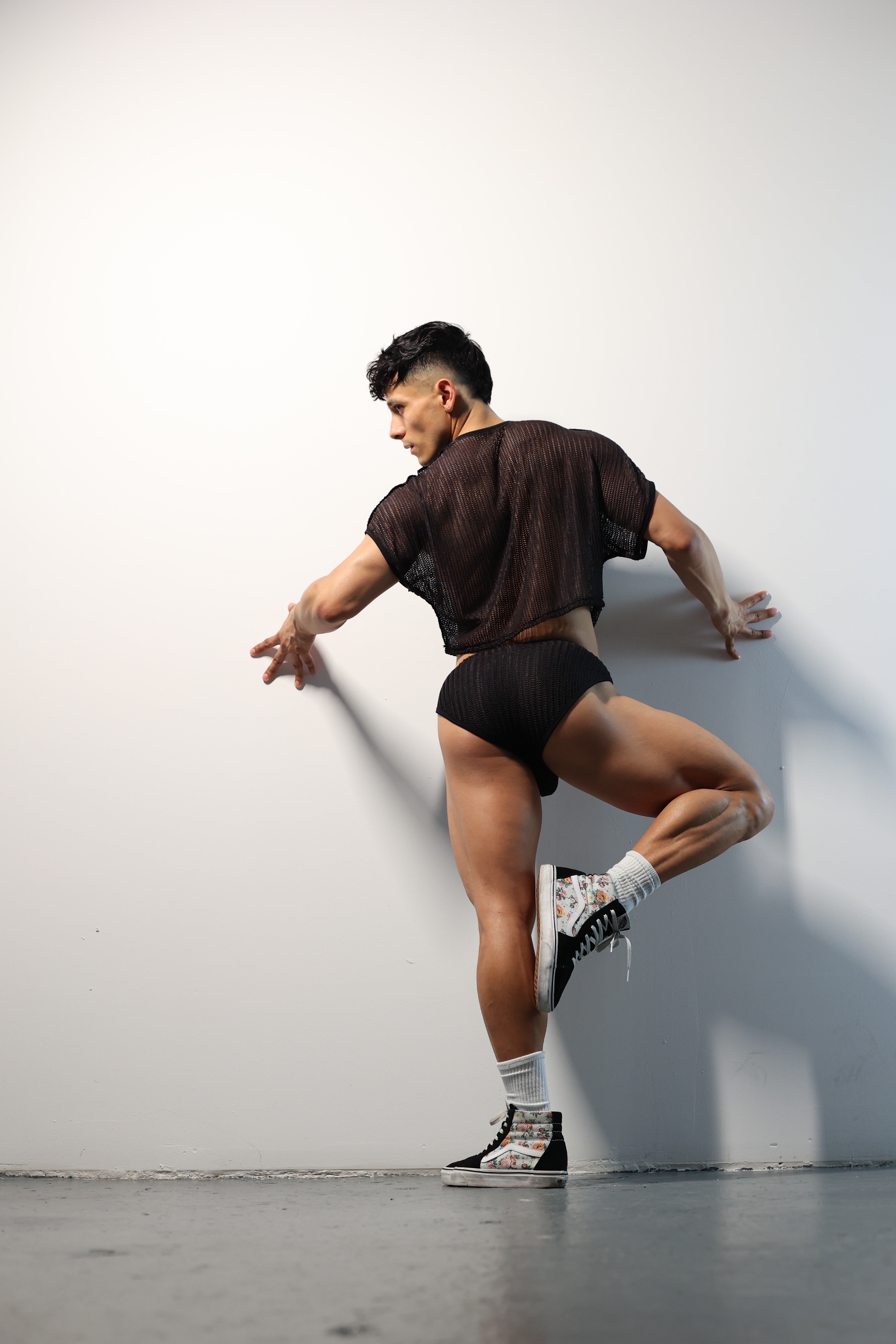 Fabian Divani modeling a sheer black mesh crop top and swim brief set—handmade by Fabi —posed in front of a white wall