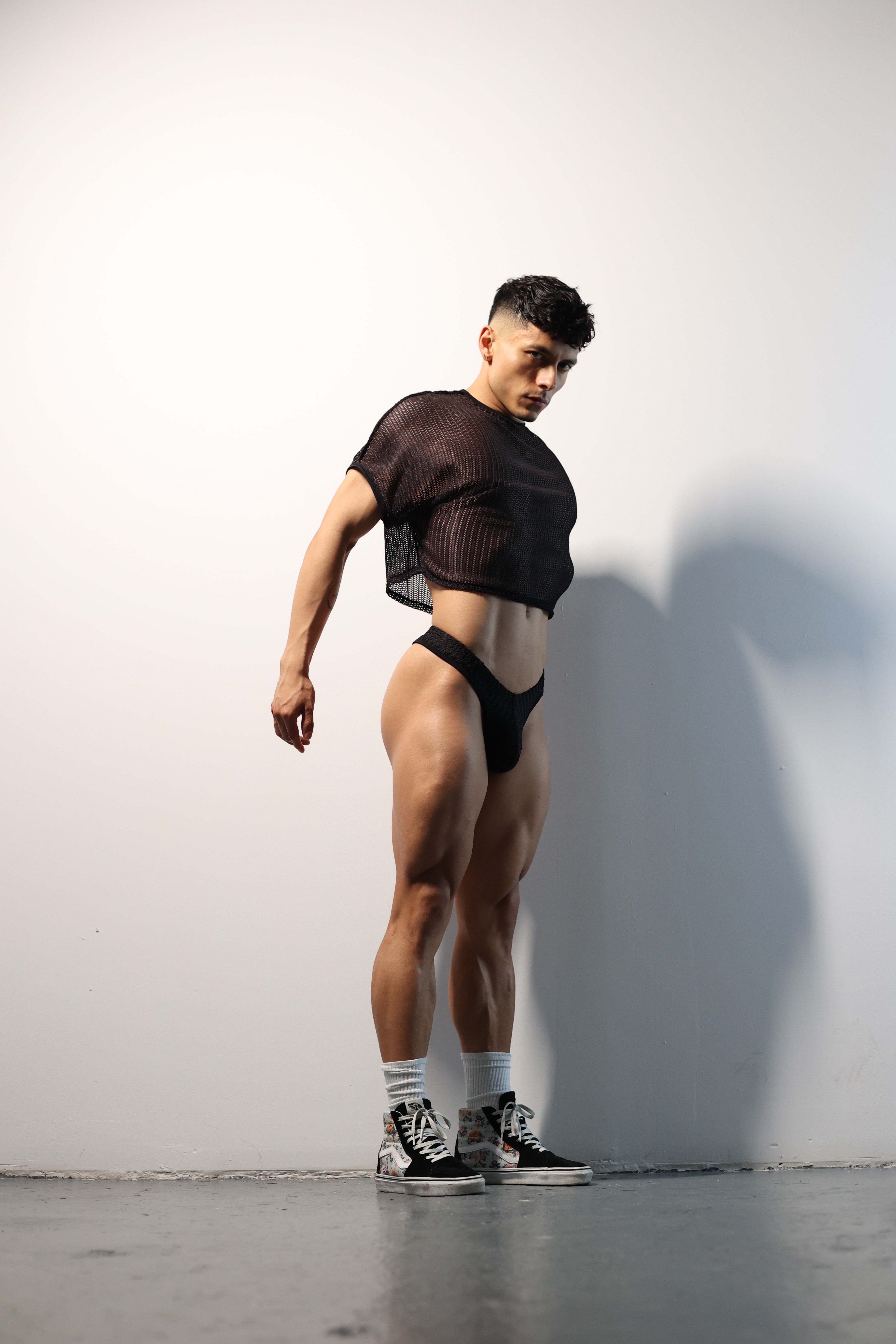 Fabian Divani modeling a sheer black mesh crop top and swim brief set—handmade by Fabi —posed in front of a white wall