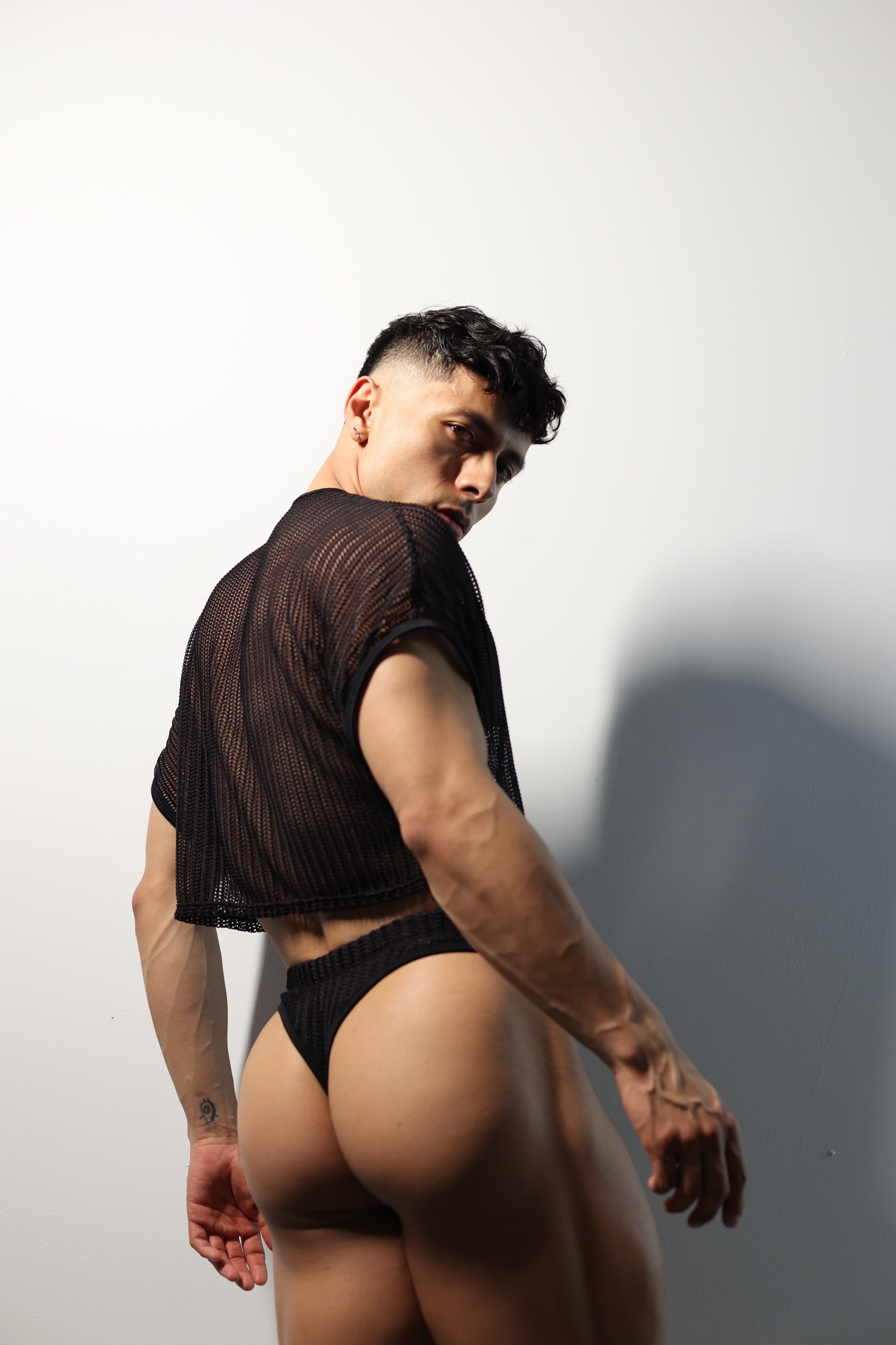 Fabian Divani modeling a sheer black mesh crop top and swim brief set—handmade by Fabi —posed in front of a white wall