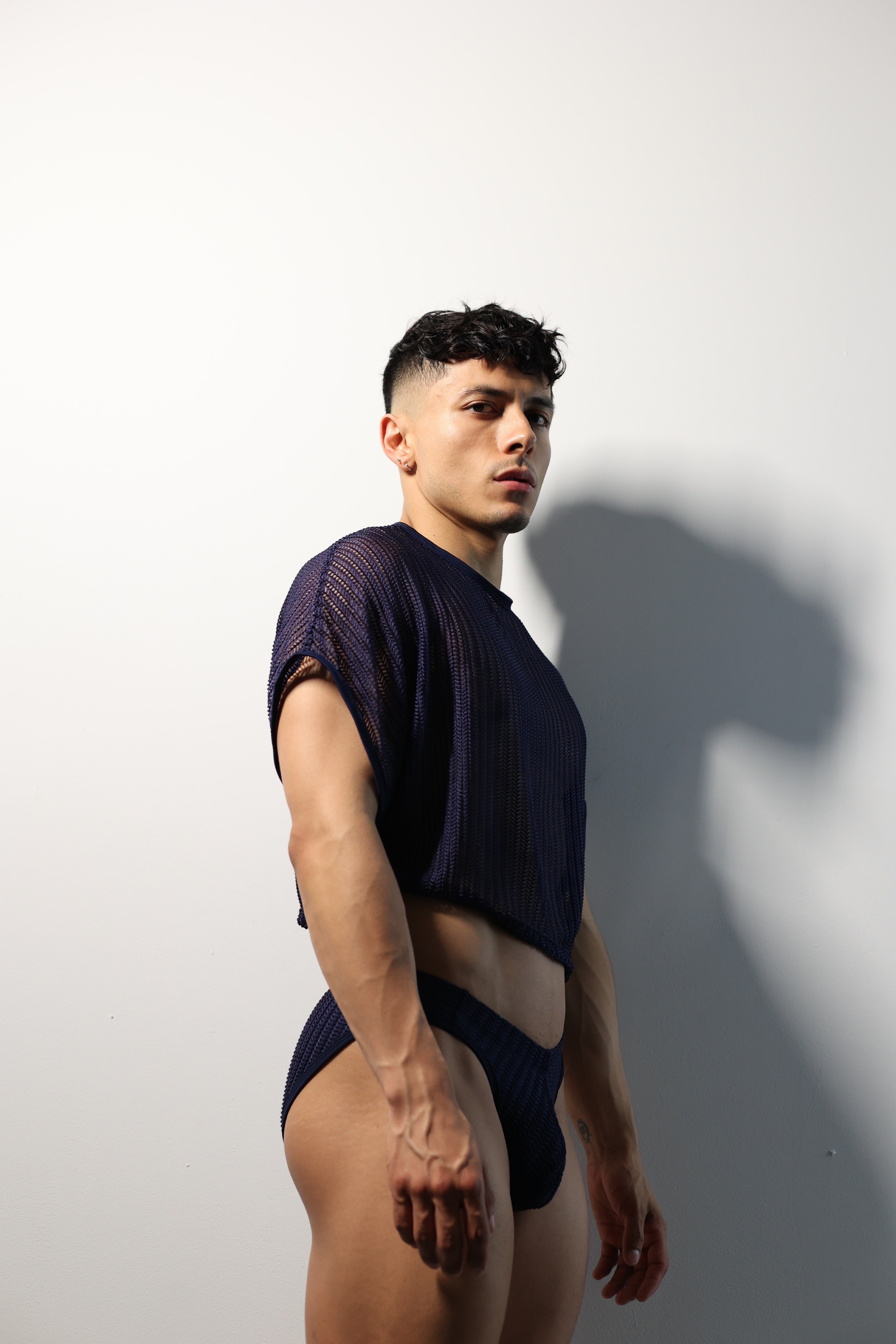Fabian Divani modeling a sheer midnight blue mesh crop top and swim brief set—handmade men's swimwear—posed in front of a white wall