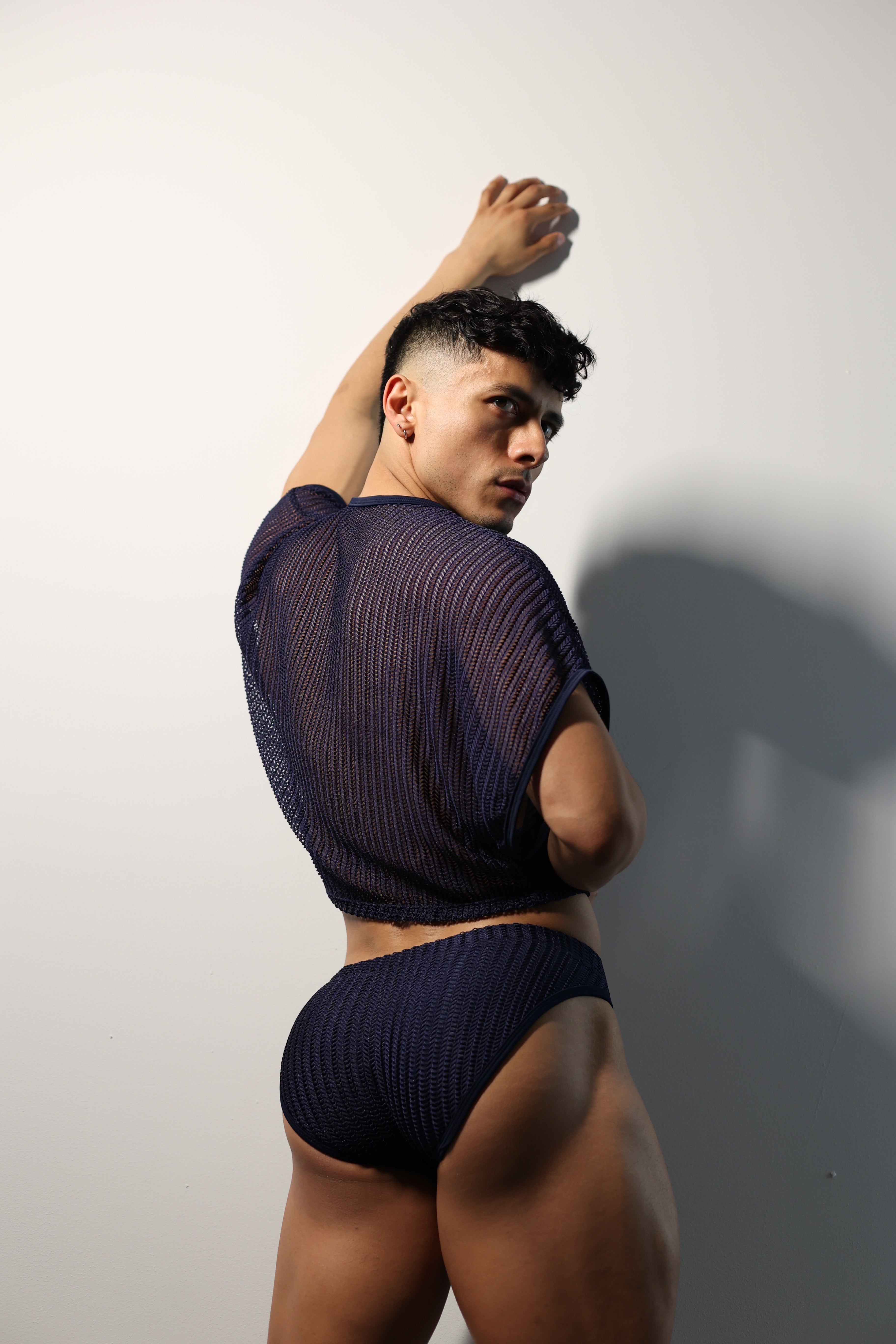Fabian Divani modeling a sheer midnight blue mesh crop top and swim brief set—handmade men's swimwear—posed in front of a white brick wall