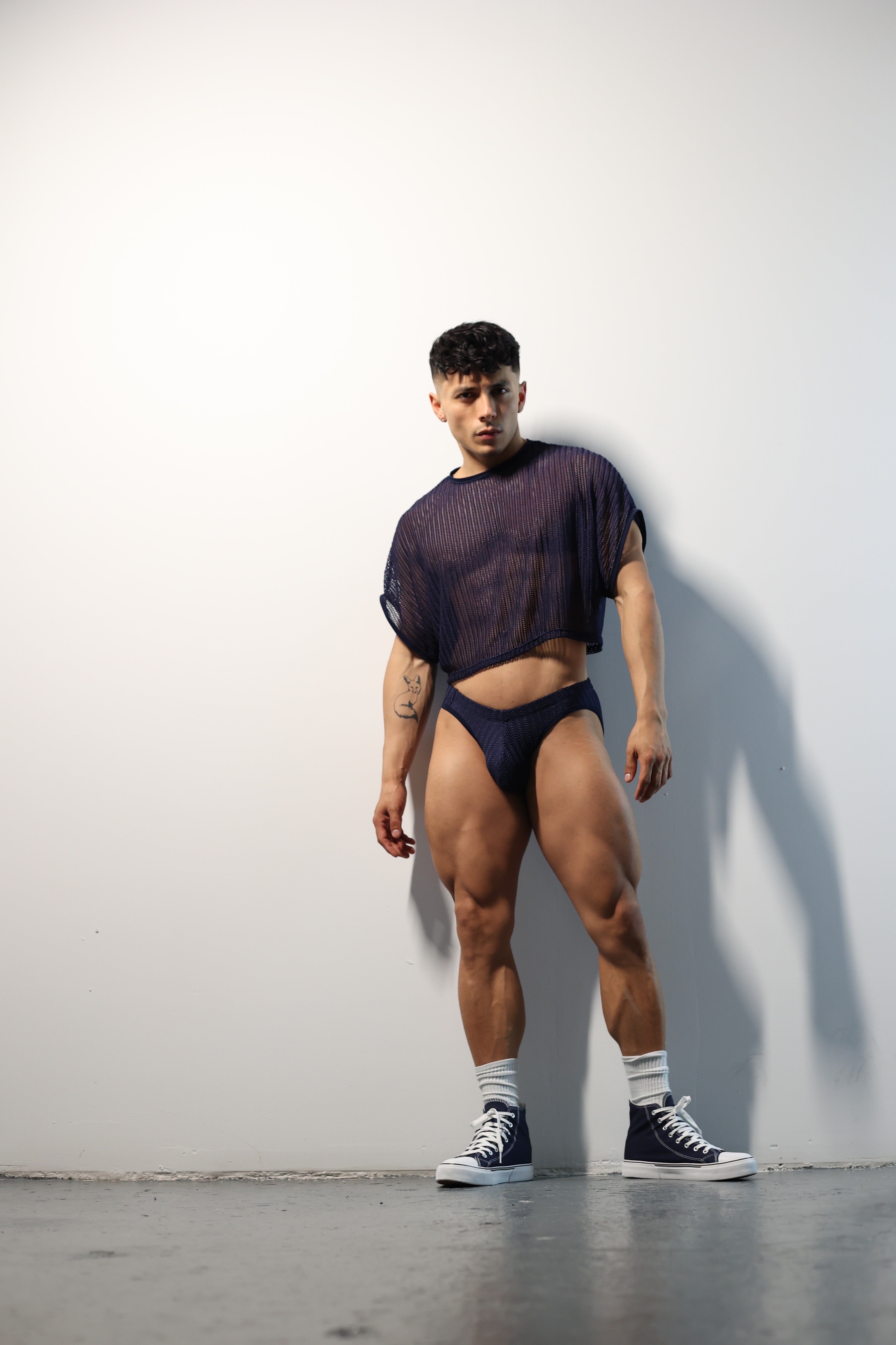 Fabian Divani modeling a sheer midnight blue mesh crop top and swim brief set—handmade men's swimwear—posed in front of a white brick wall