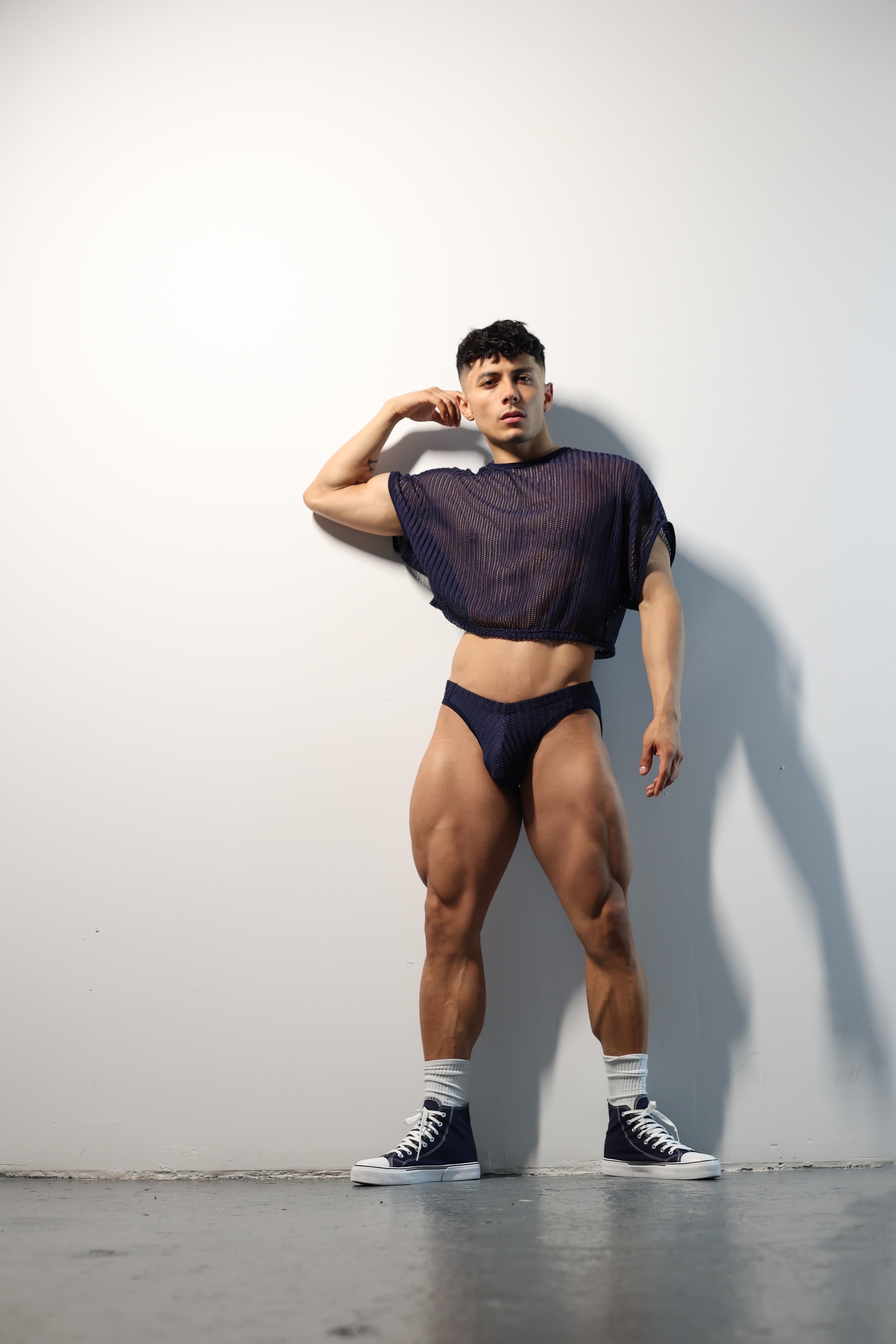 Fabian Divani modeling a sheer midnight blue mesh crop top and swim brief set—handmade men's swimwear—posed in front of a white brick wall
