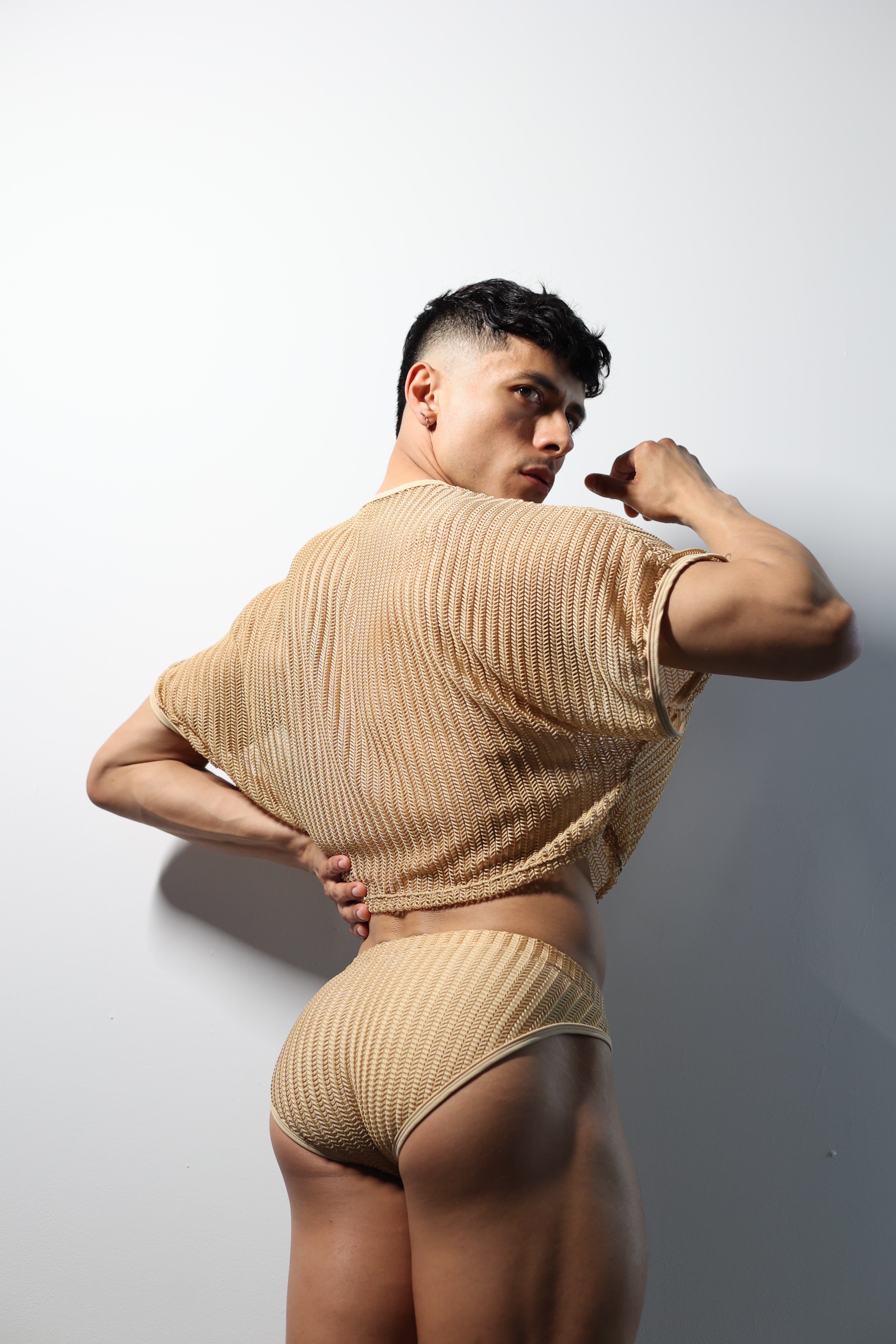 Fabian Divani modeling a sheer natural mesh crop top and swim brief set—handmade men's swimwear—posed in front of a white wall