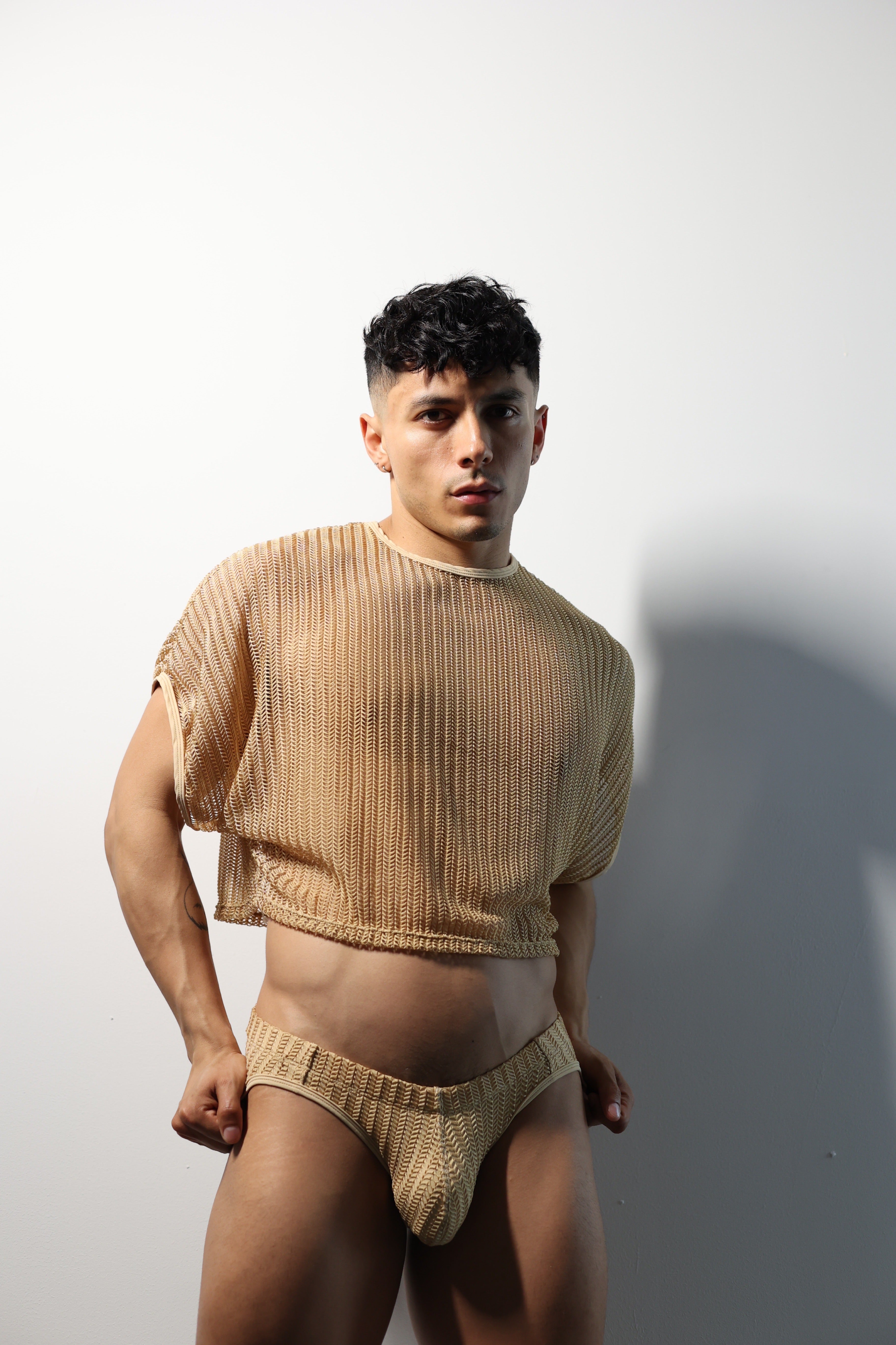 Fabian Divani modeling a sheer natural mesh crop top and swim brief set—handmade men's swimwear—posed in front of a white wall