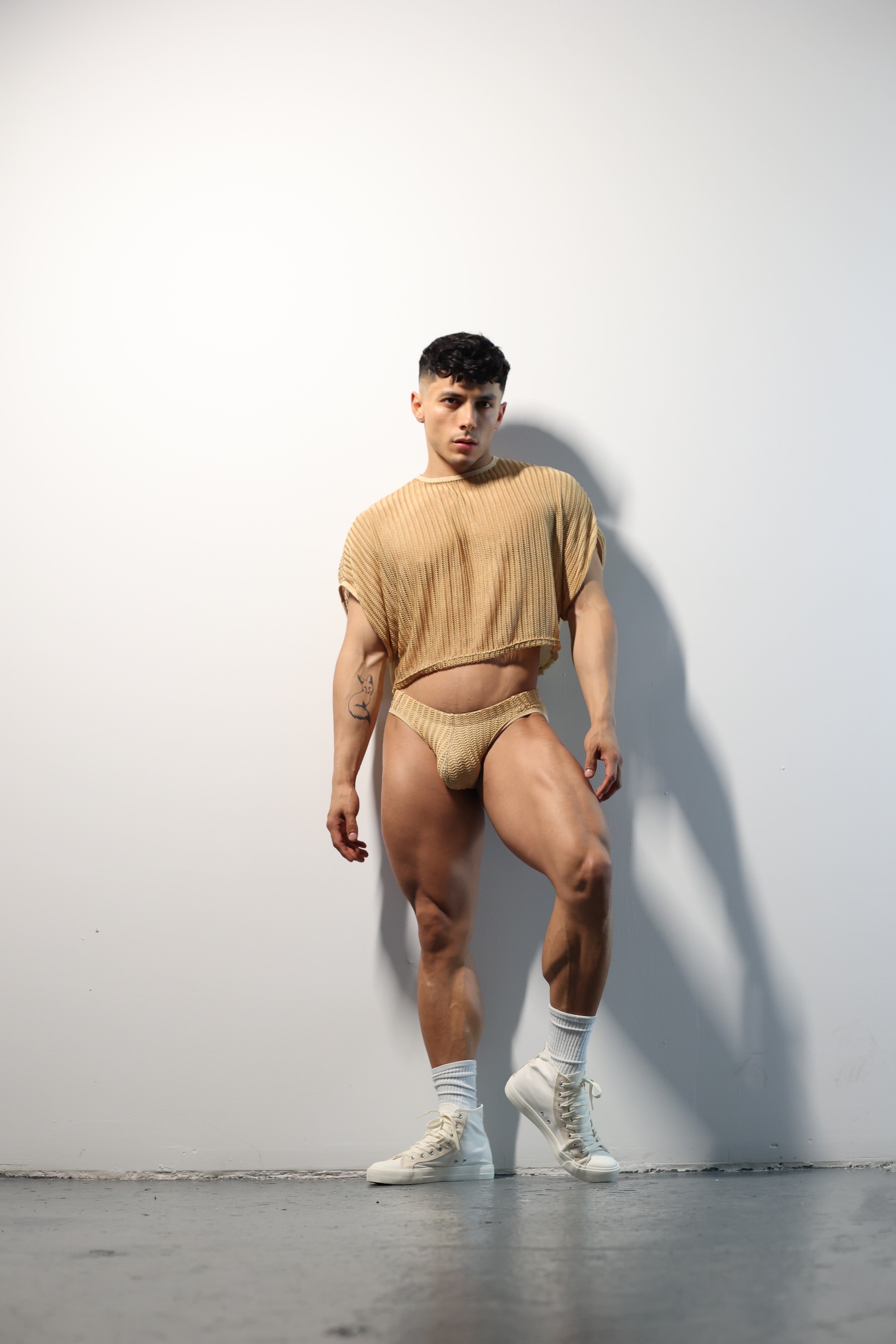 Fabian Divani modeling a sheer natural mesh crop top and swim brief set—handmade men's swimwear—posed in front of a white wall