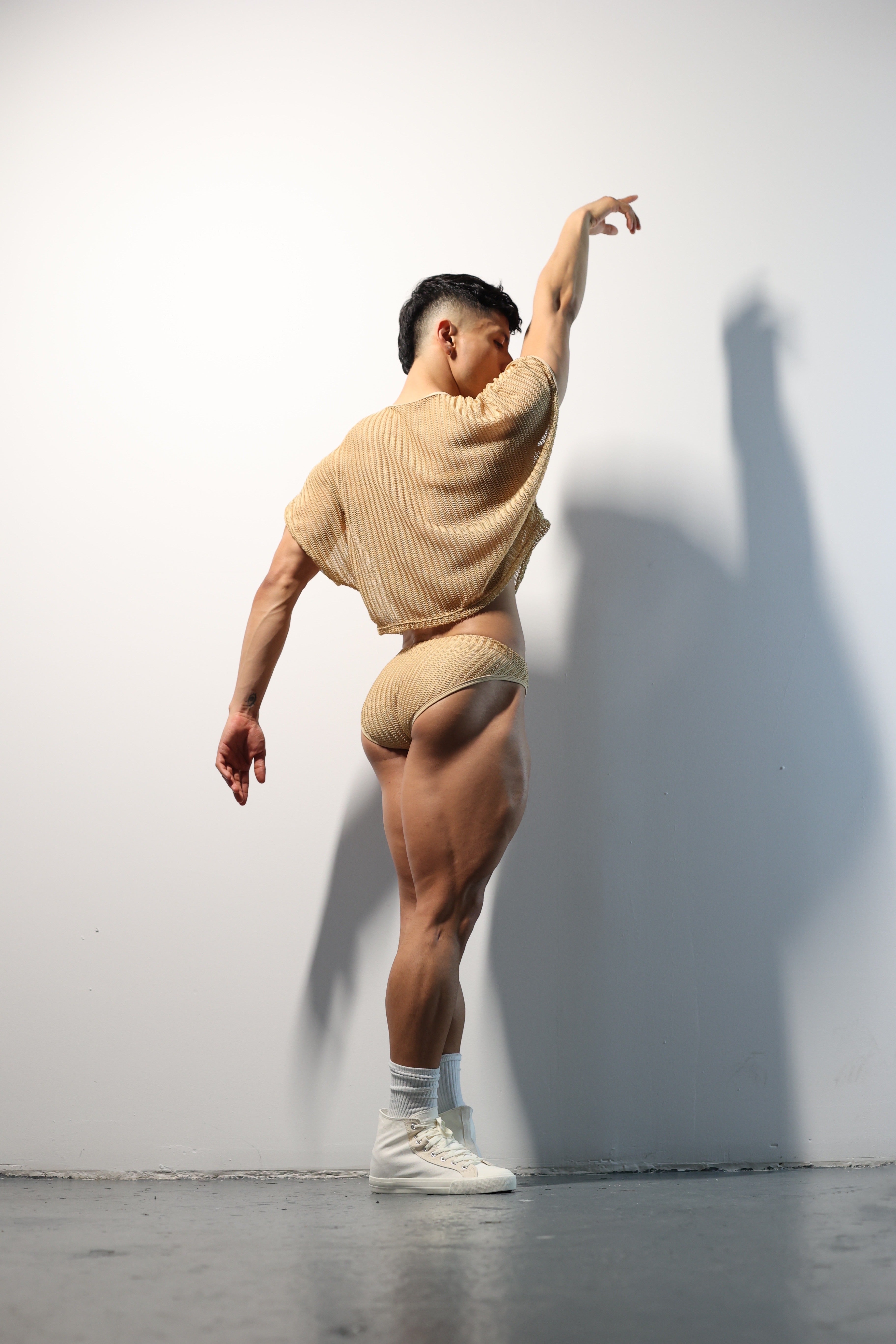 Fabian Divani modeling a sheer natural mesh crop top and swim brief set—handmade men's swimwear—posed in front of a white wall