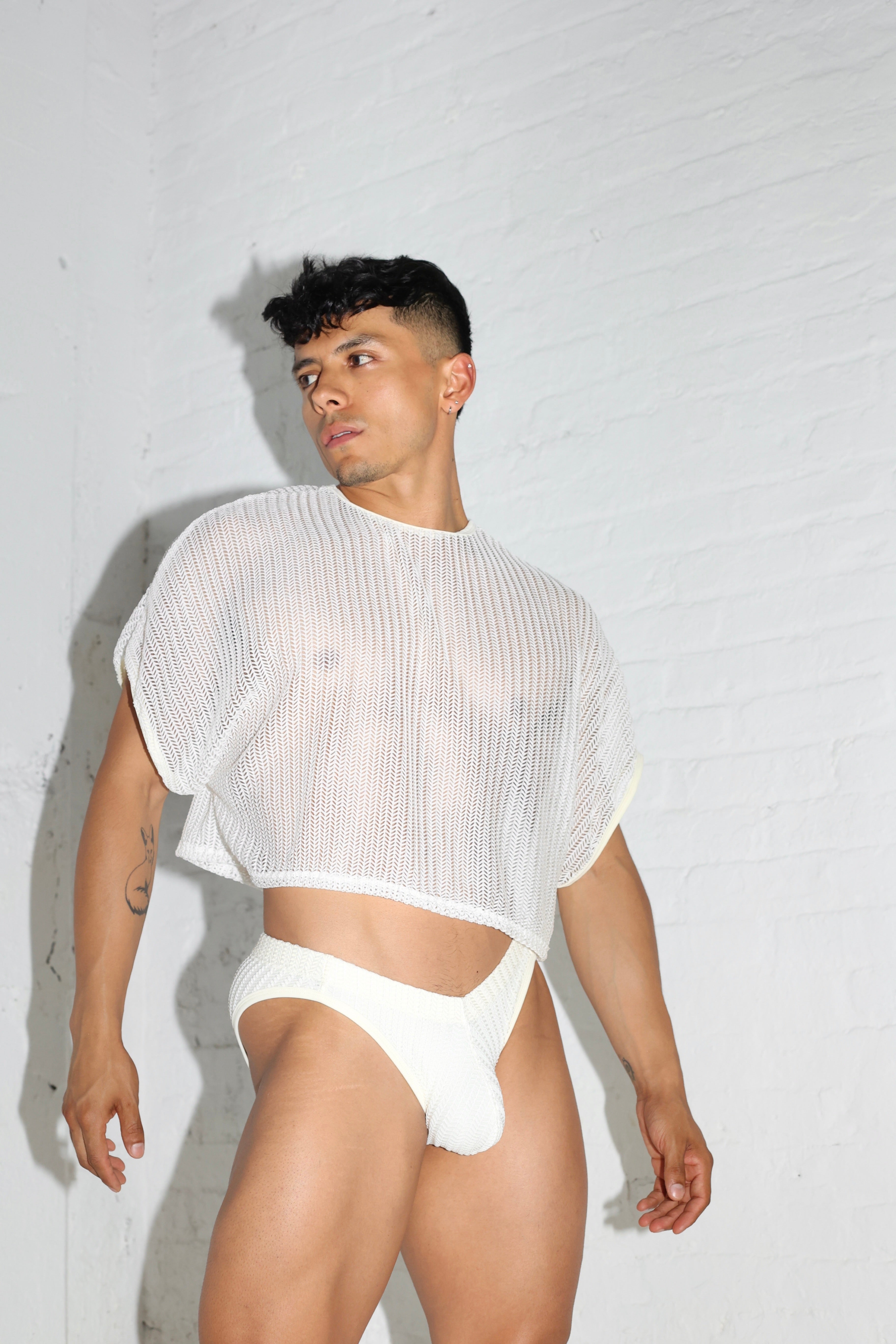Fabian Divani modeling a sheer white mesh crop top and swim brief set—handmade by Fabi —posed in front of a white brick wall