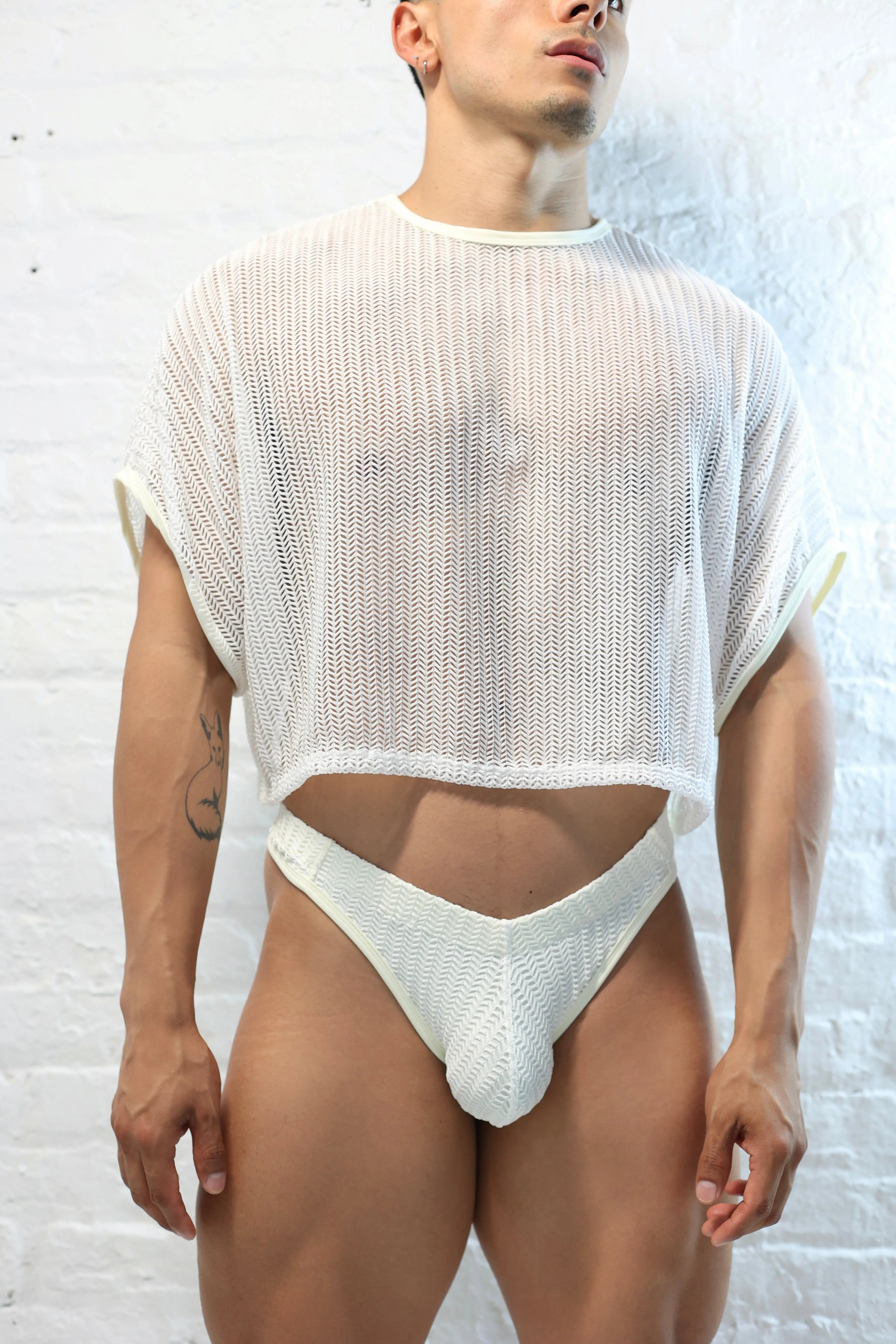 Fabian Divani modeling a sheer white mesh crop top and swim thong set—handmade men's swimwear—posed in front of a white brick wall