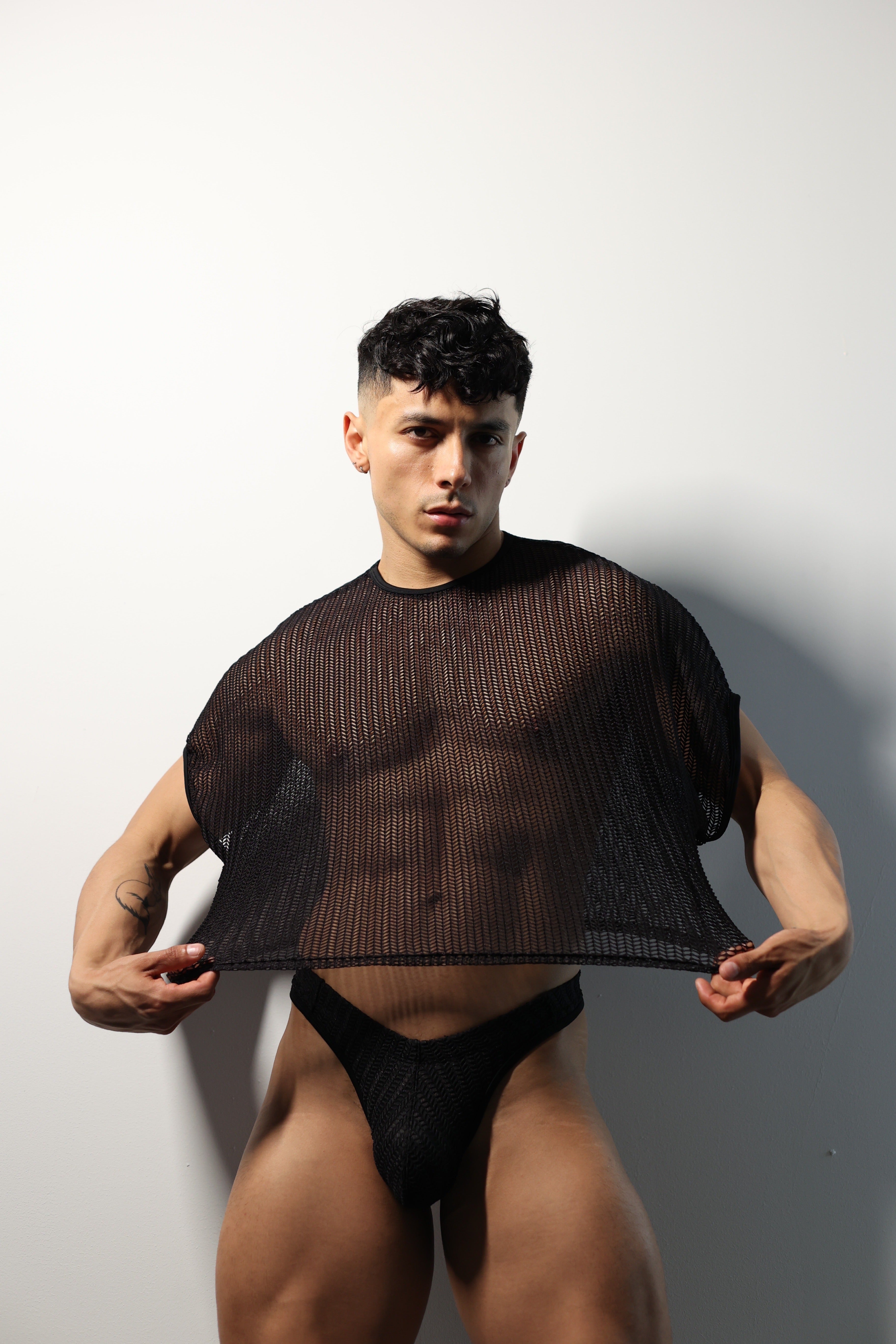 Fabian Divani modeling a sheer black mesh crop top and swim brief set—handmade by Fabi —posed in front of a white brick wall