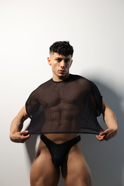 Fabian Divani modeling a sheer black mesh crop top and swim brief set—handmade by Fabi —posed in front of a white brick wall