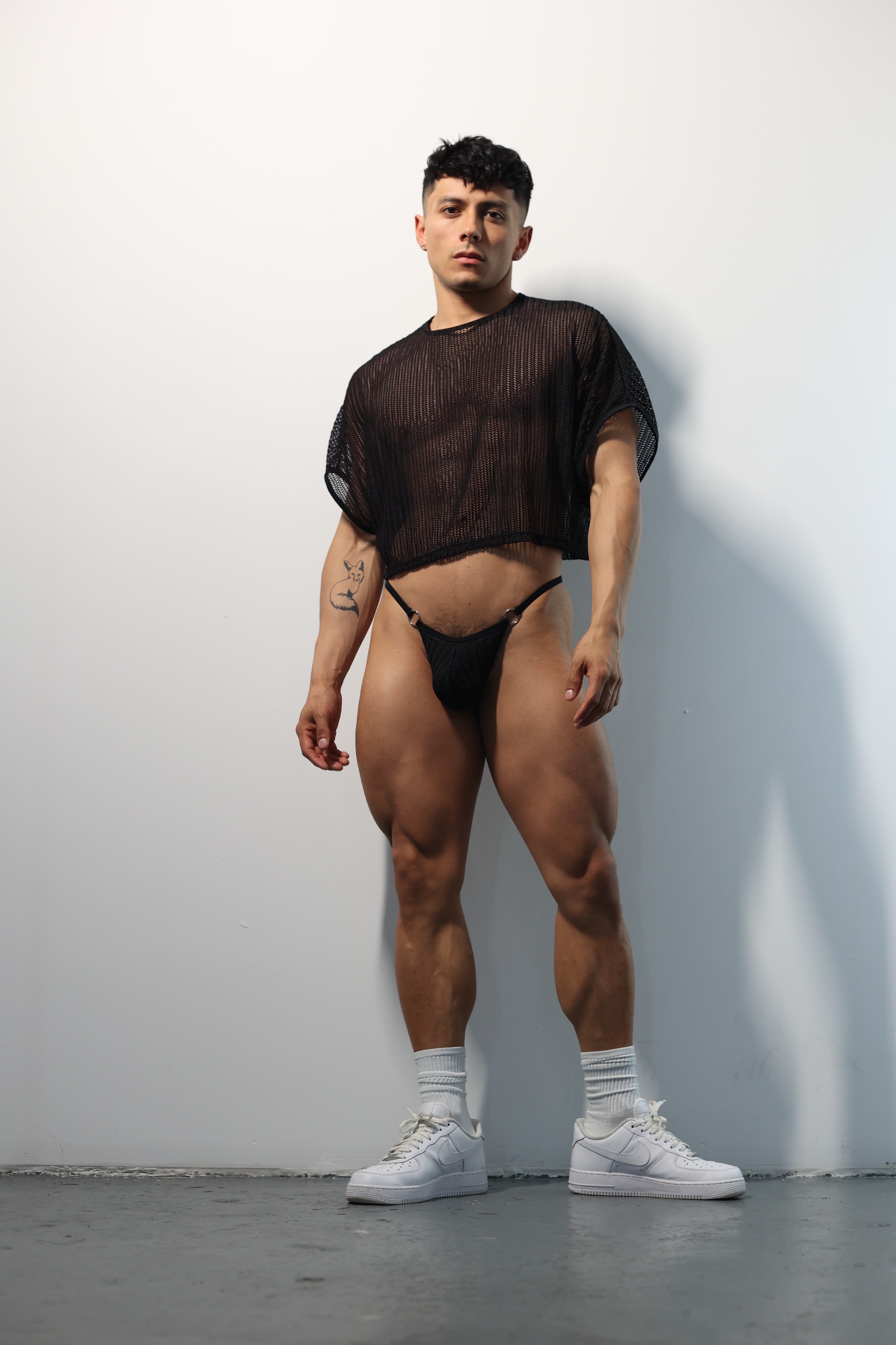 Fabian Divani modeling a sheer black mesh crop top and G-string underwear set— handmade men's underwear — posed in front of a white brick wall