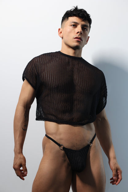 Fabian Divani modeling a sheer black mesh crop top and G-string underwear set— handmade men's underwear — posed in front of a white brick wall