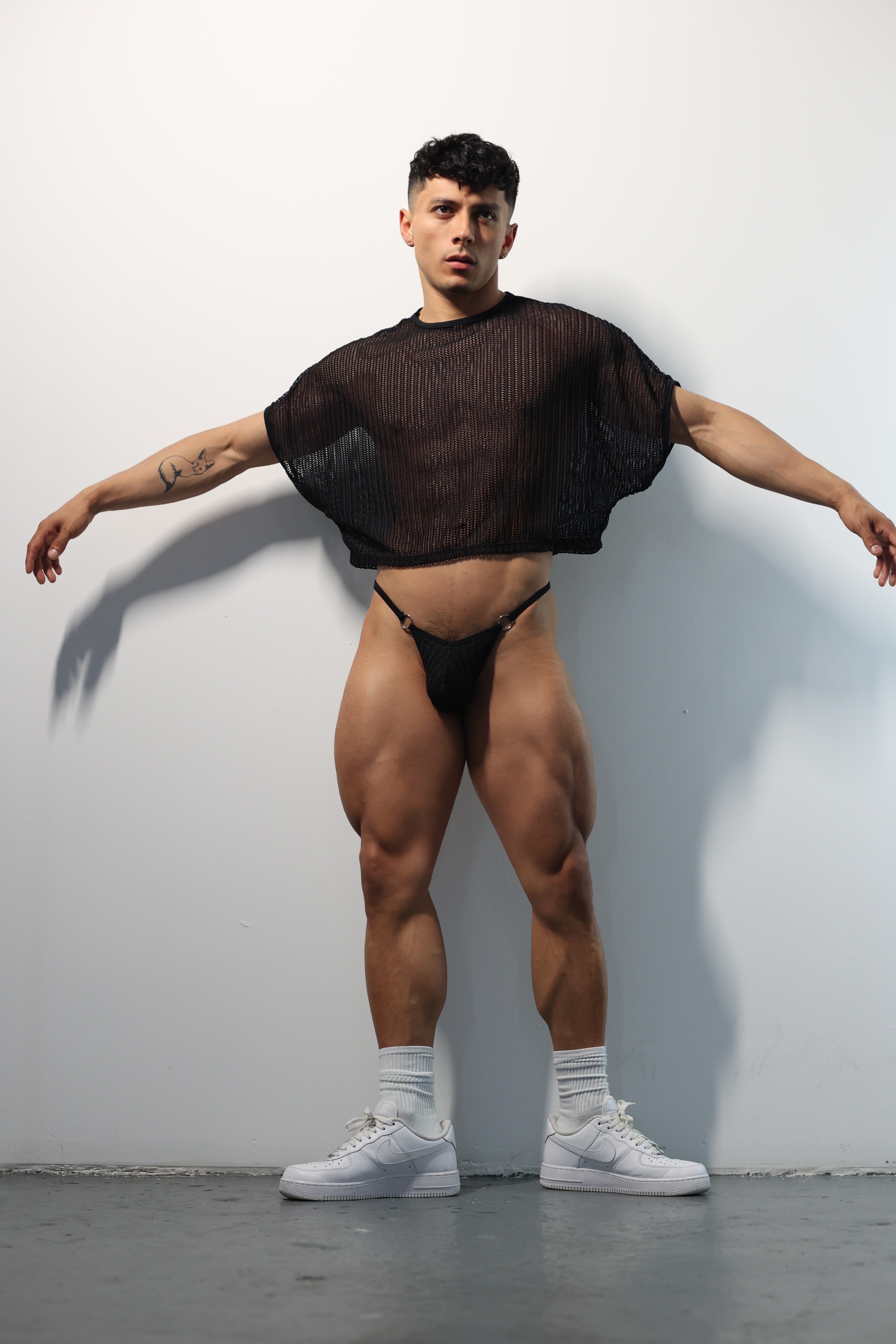 Fabian Divani modeling a sheer black mesh crop top and G-string underwear set— handmade men's underwear — posed in front of a white brick wall