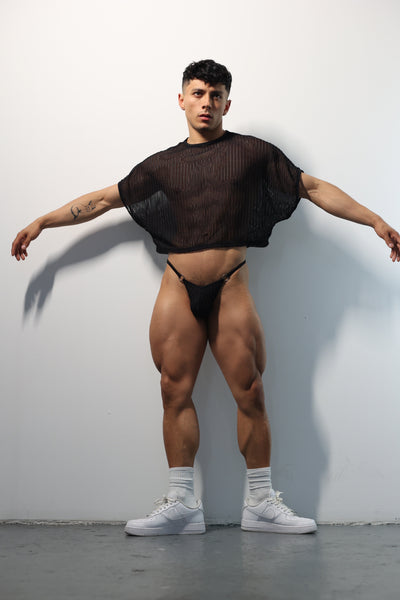 Fabian Divani modeling a sheer black mesh crop top and G-string underwear set— handmade men's underwear — posed in front of a white brick wall