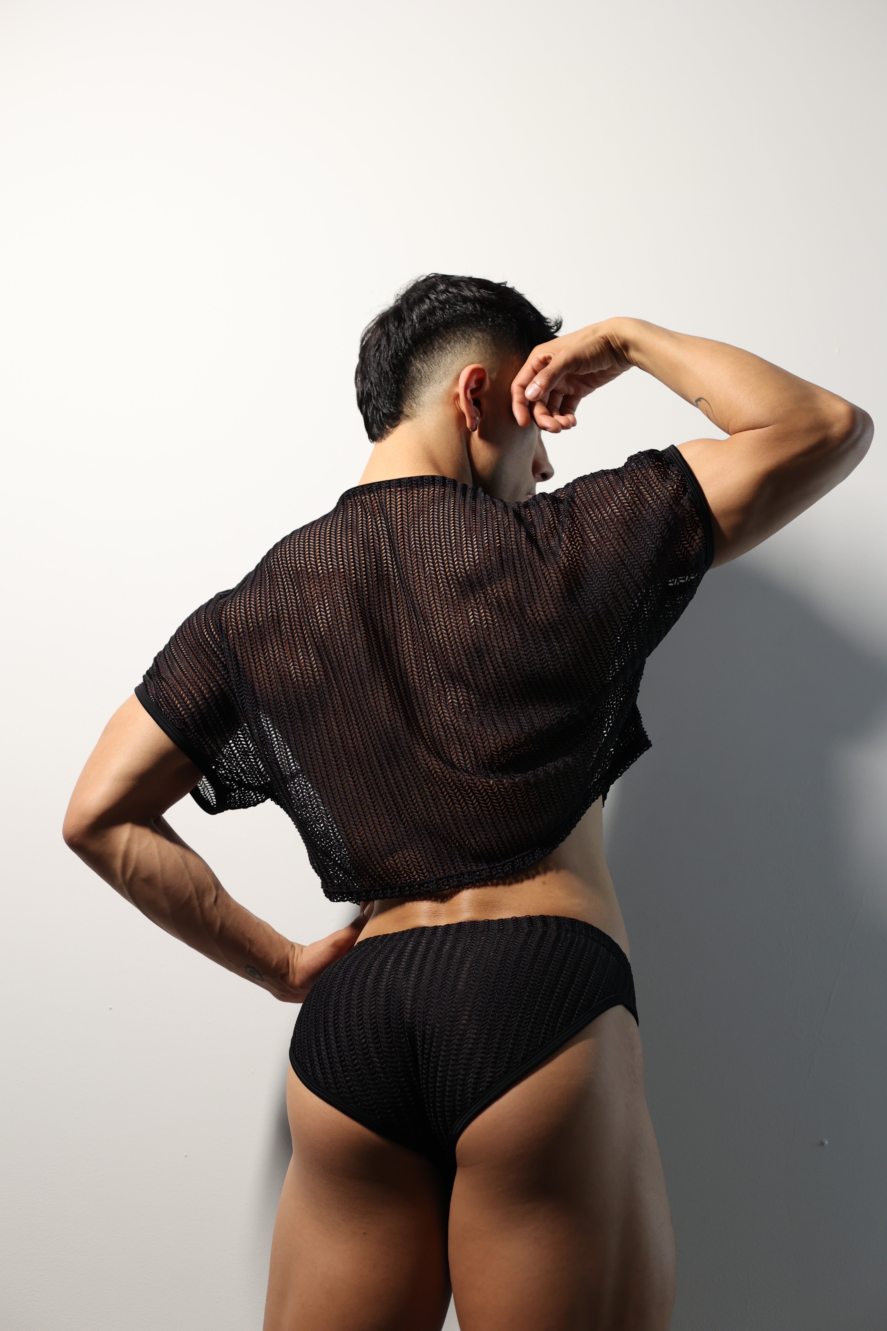 Fabian Divani modeling a sheer black mesh crop top and swim brief set—handmade by Fabi —posed in front of a white wall