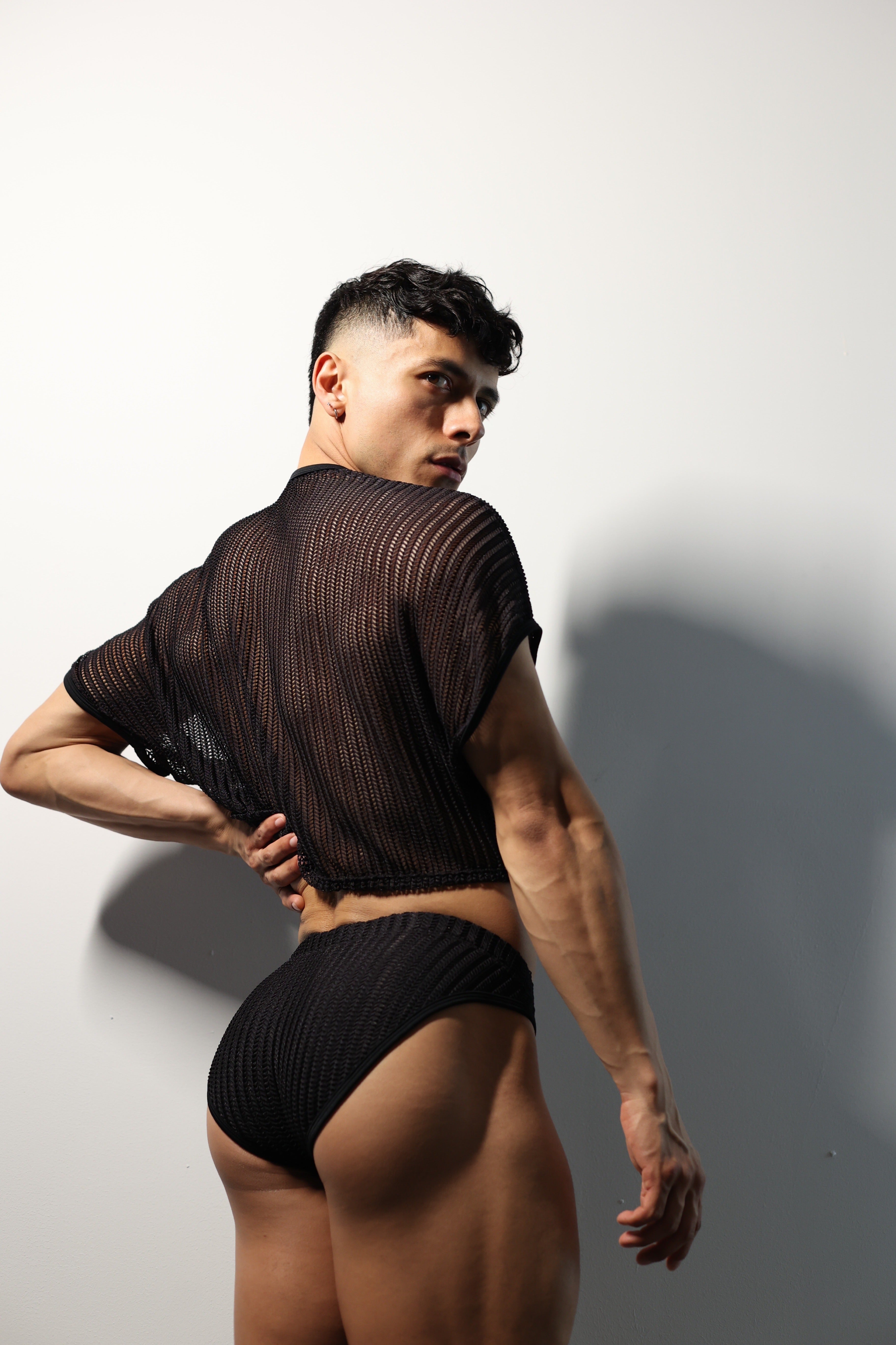 Fabian Divani modeling a sheer black mesh crop top and swim brief set—handmade by Fabi —posed in front of a white wall