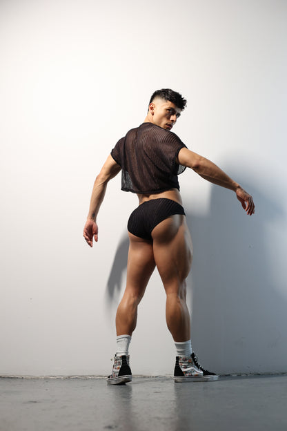 Fabian Divani modeling a sheer black mesh crop top and swim brief set—handmade by Fabi —posed in front of a white wall