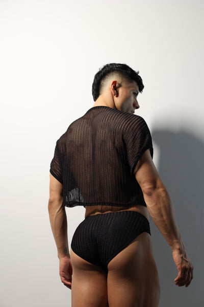Fabian Divani modeling a sheer black mesh crop top and swim brief set—handmade by Fabi —posed in front of a white wall
