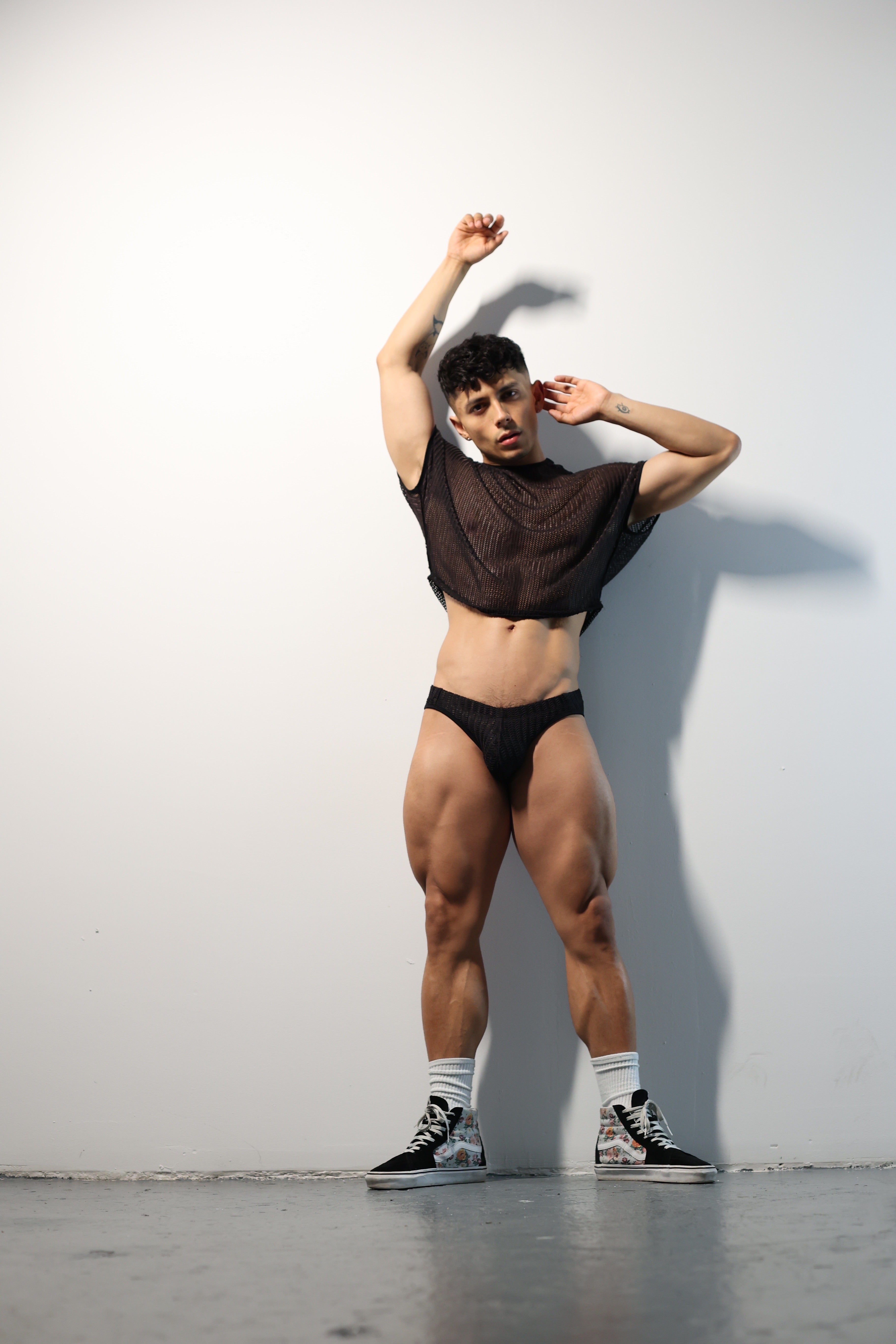 Fabian Divani modeling a sheer black mesh crop top and swim brief set—handmade by Fabi —posed in front of a white wall