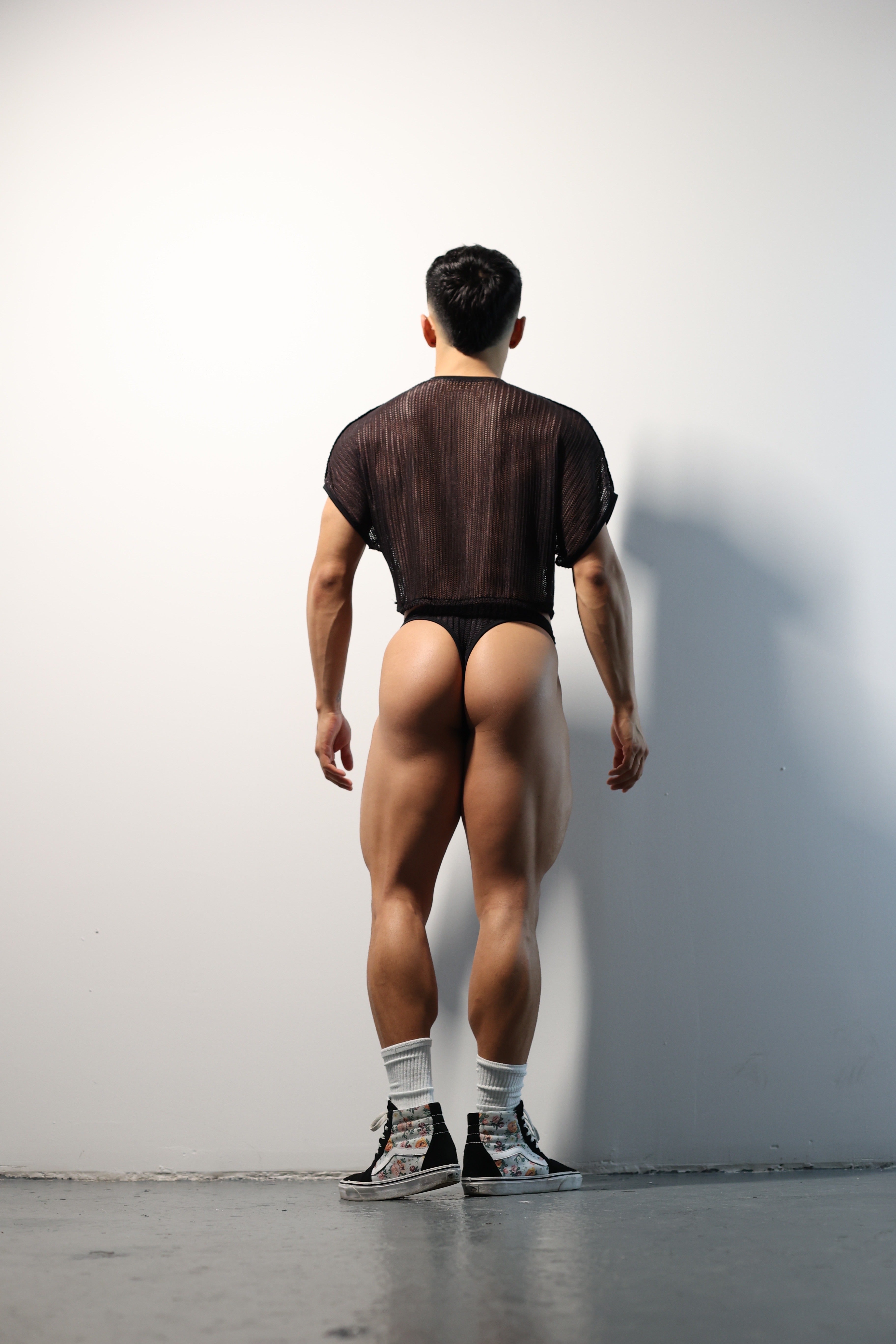 Fabian Divani modeling a sheer black mesh crop top and swim brief set—handmade by Fabi —posed in front of a white brick wall
