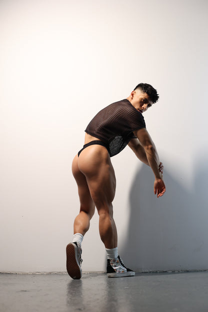 Fabian Divani modeling a sheer black mesh crop top and swim brief set—handmade by Fabi —posed in front of a white wall