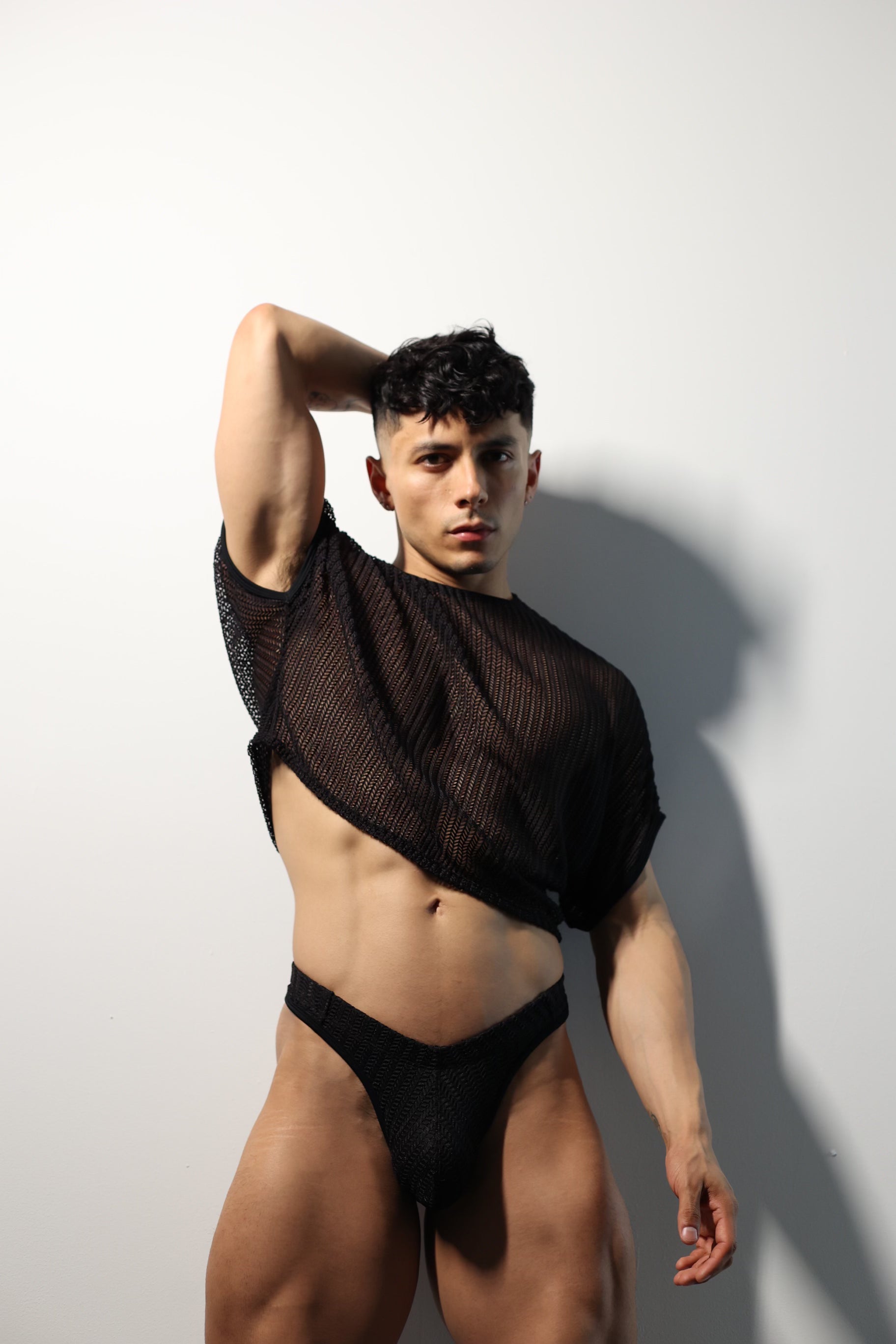 Fabian Divani modeling a sheer black mesh crop top and swim brief set—handmade by Fabi —posed in front of a white brick wall