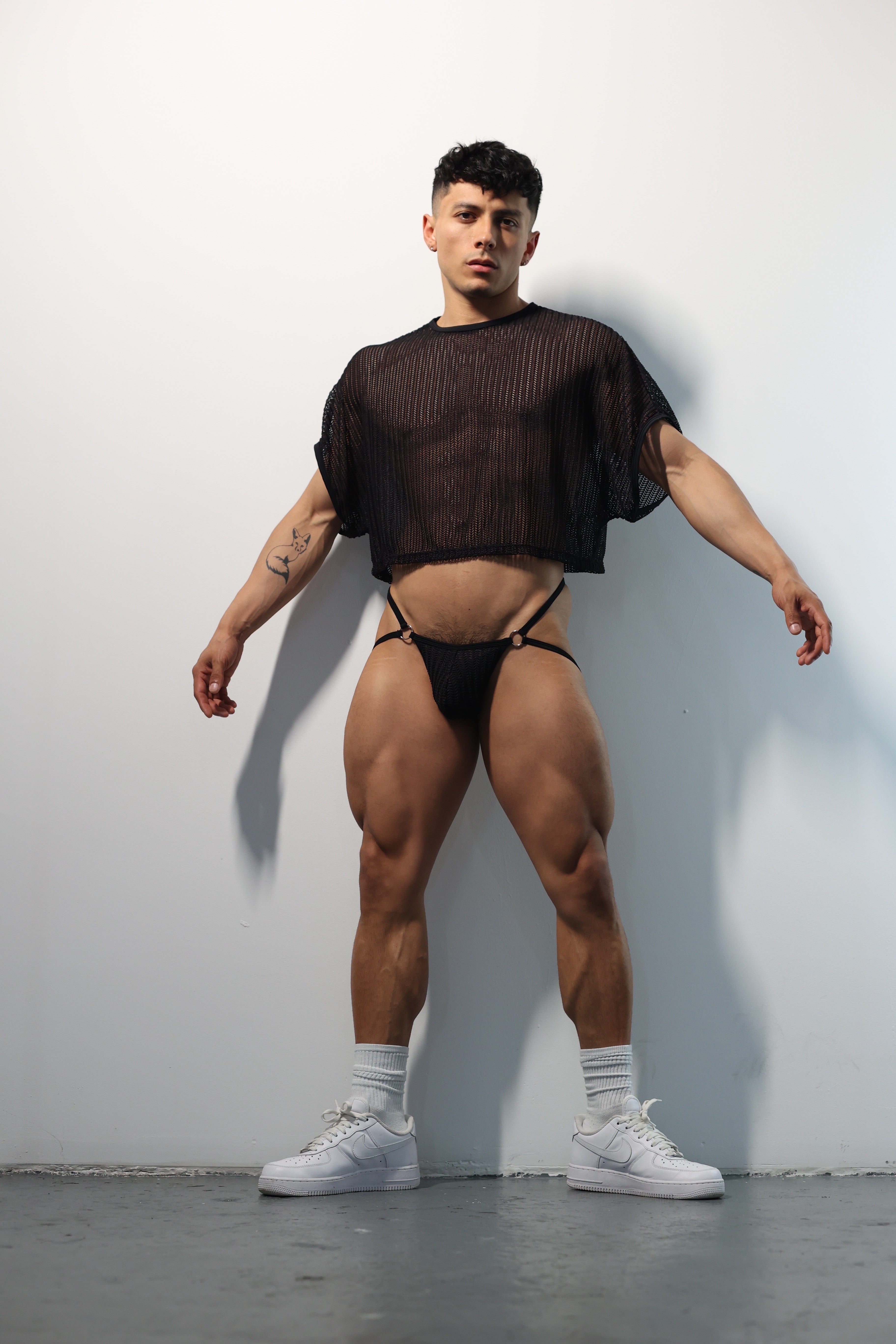 Fabian Divani modeling a sheer black mesh crop top and string jockstrap set—handmade men's underwear—posed in front of a white wall