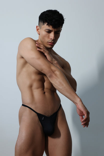 Fabian Divani modeling a black ribbed G-string underwear set— handmade men's underwear — posed in front of a white brick wall
