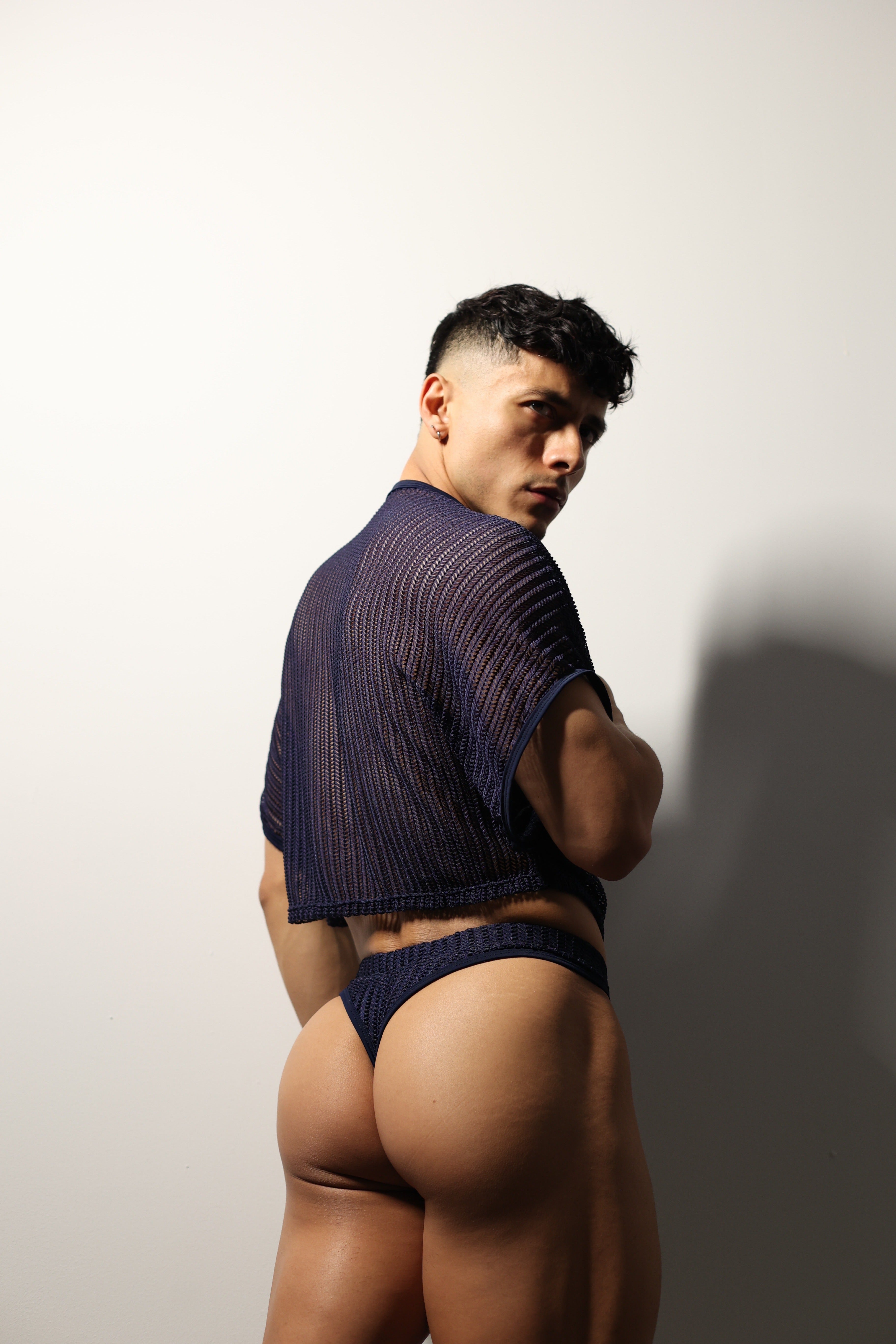 Fabian Divani modeling a sheer midnight blue mesh crop top and swim brief set—handmade men's swimwear—posed in front of a white wall