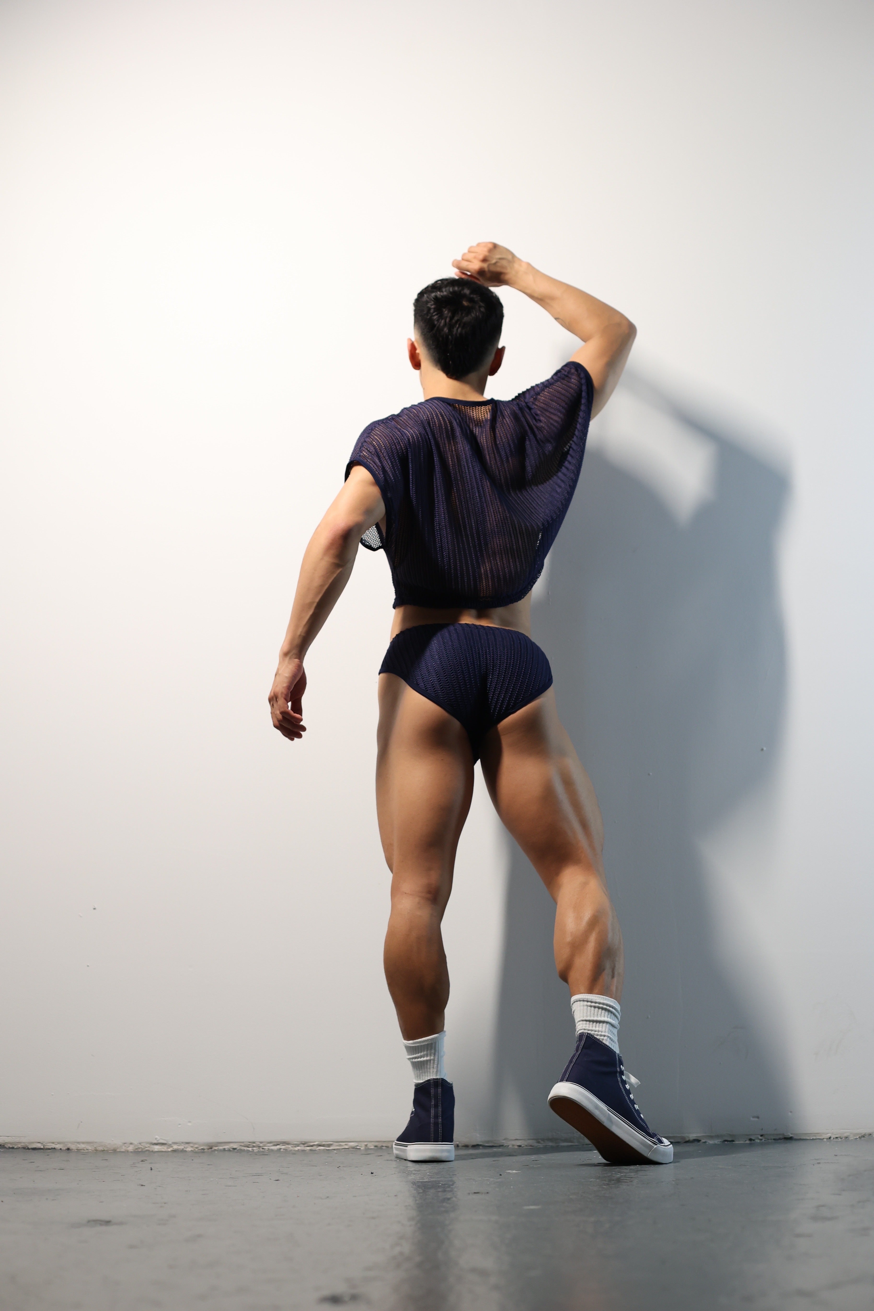 Fabian Divani modeling a sheer midnight blue mesh crop top and swim brief set—handmade men's swimwear—posed in front of a white brick wall