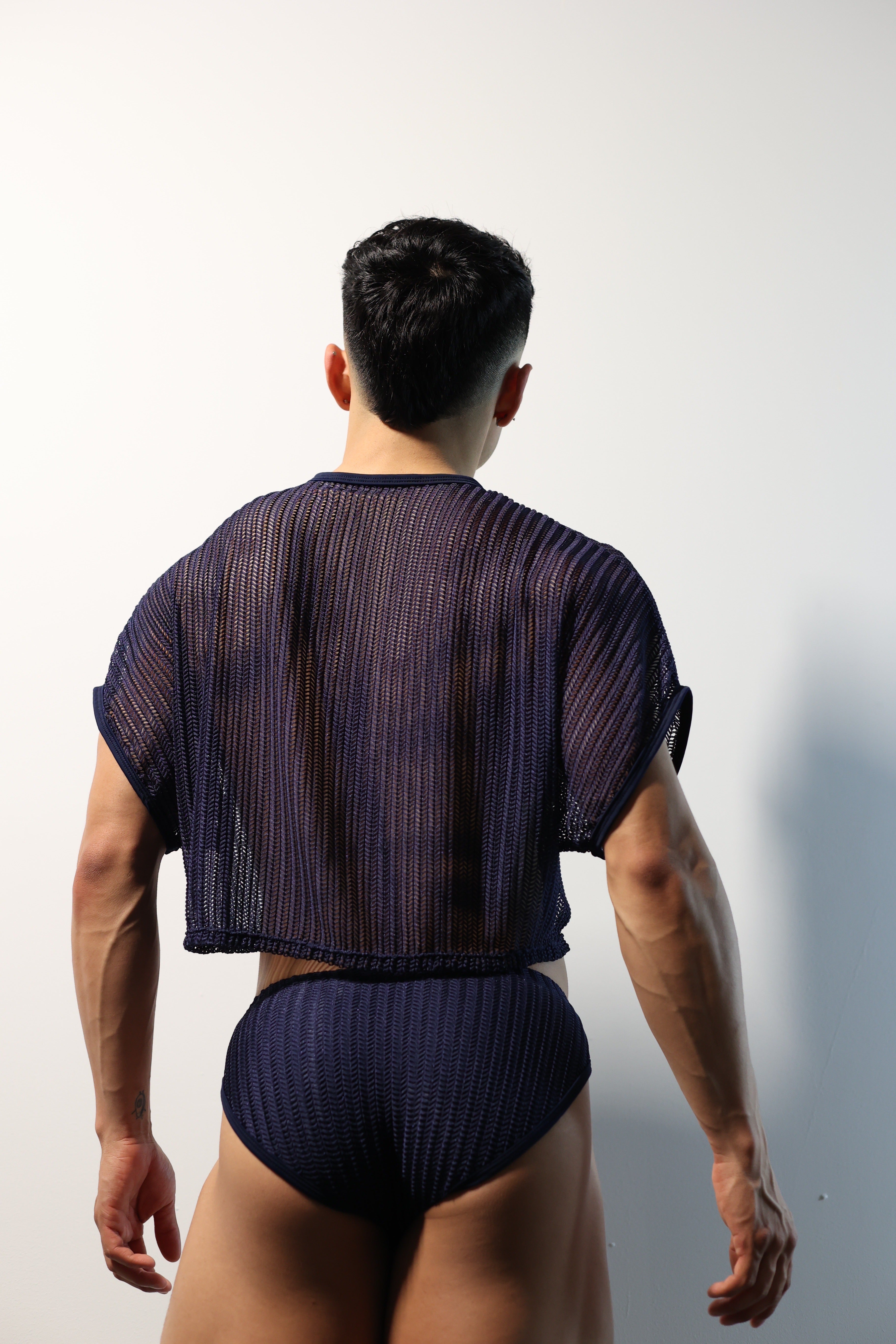 Fabian Divani modeling a sheer midnight blue mesh crop top and swim brief set—handmade men's swimwear—posed in front of a white brick wall