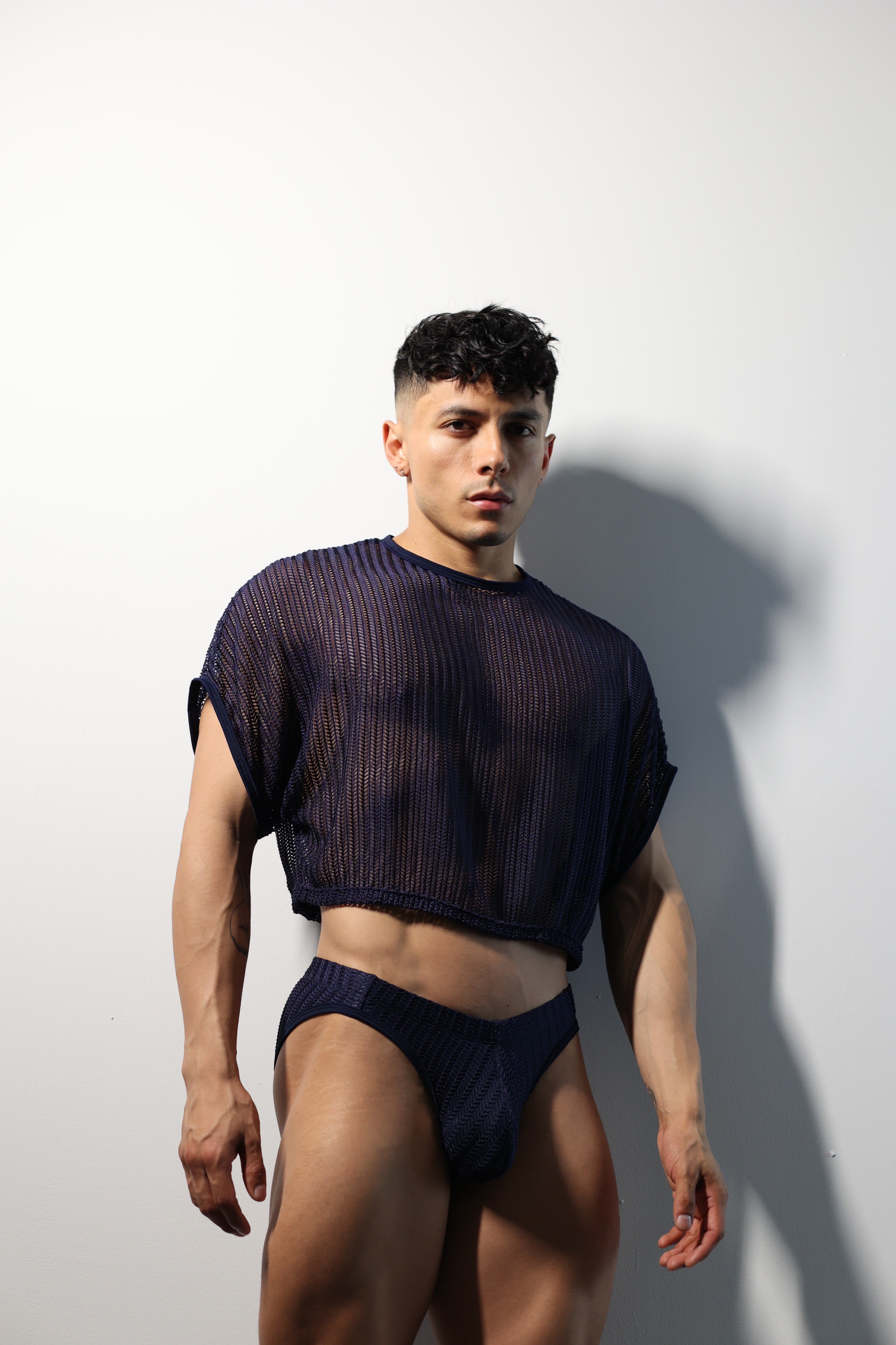 Fabian Divani modeling a sheer midnight blue mesh crop top and swim brief set—handmade men's swimwear—posed in front of a white brick wall