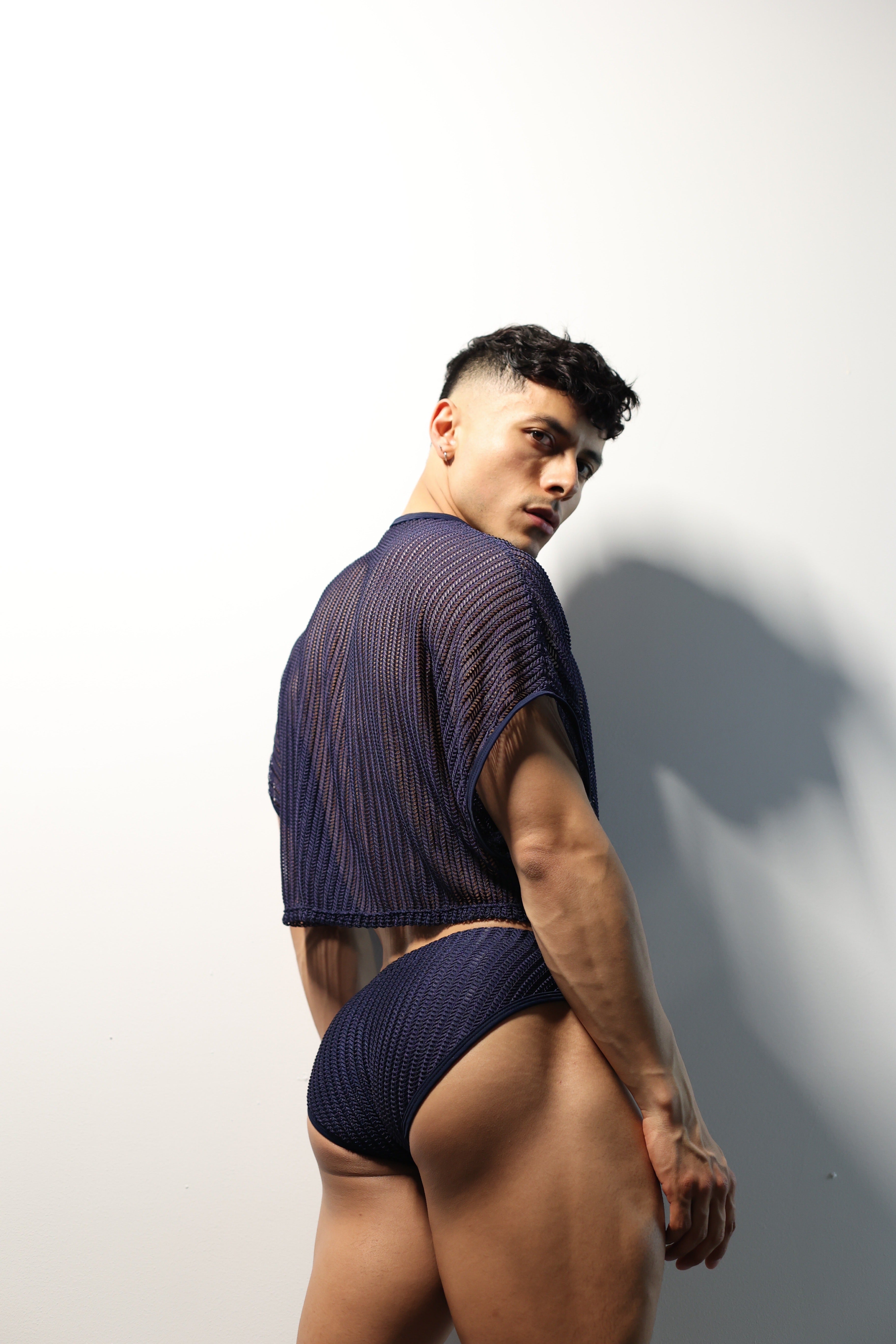 Fabian Divani modeling a sheer midnight blue mesh crop top and swim brief set—handmade men's swimwear—posed in front of a white brick wall