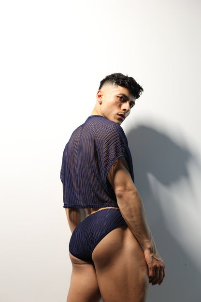 Fabian Divani modeling a sheer midnight blue mesh crop top and swim brief set—handmade men's swimwear—posed in front of a white brick wall