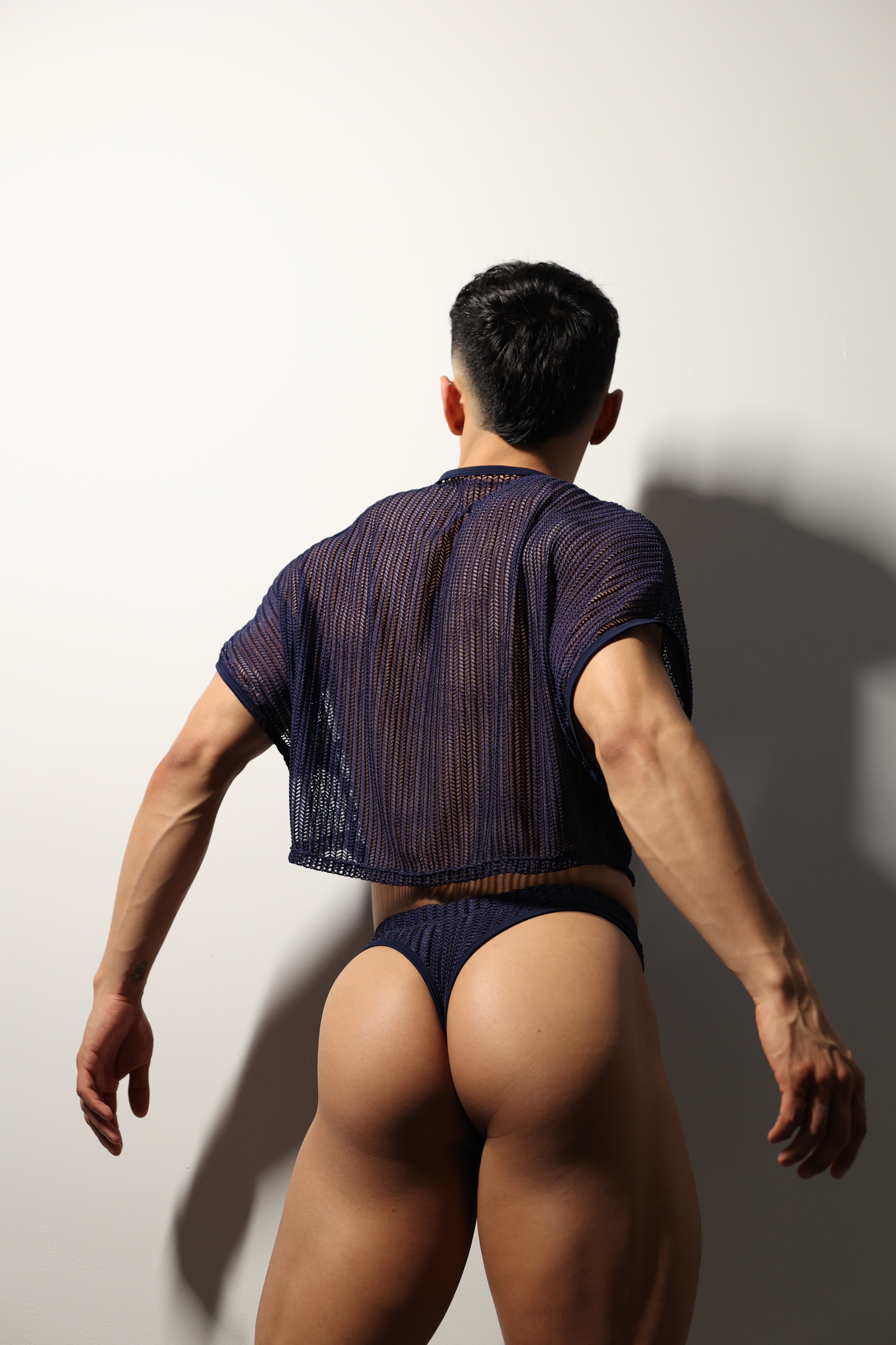 Fabian Divani modeling a sheer midnight blue mesh crop top and swim brief set—handmade men's swimwear—posed in front of a white wall