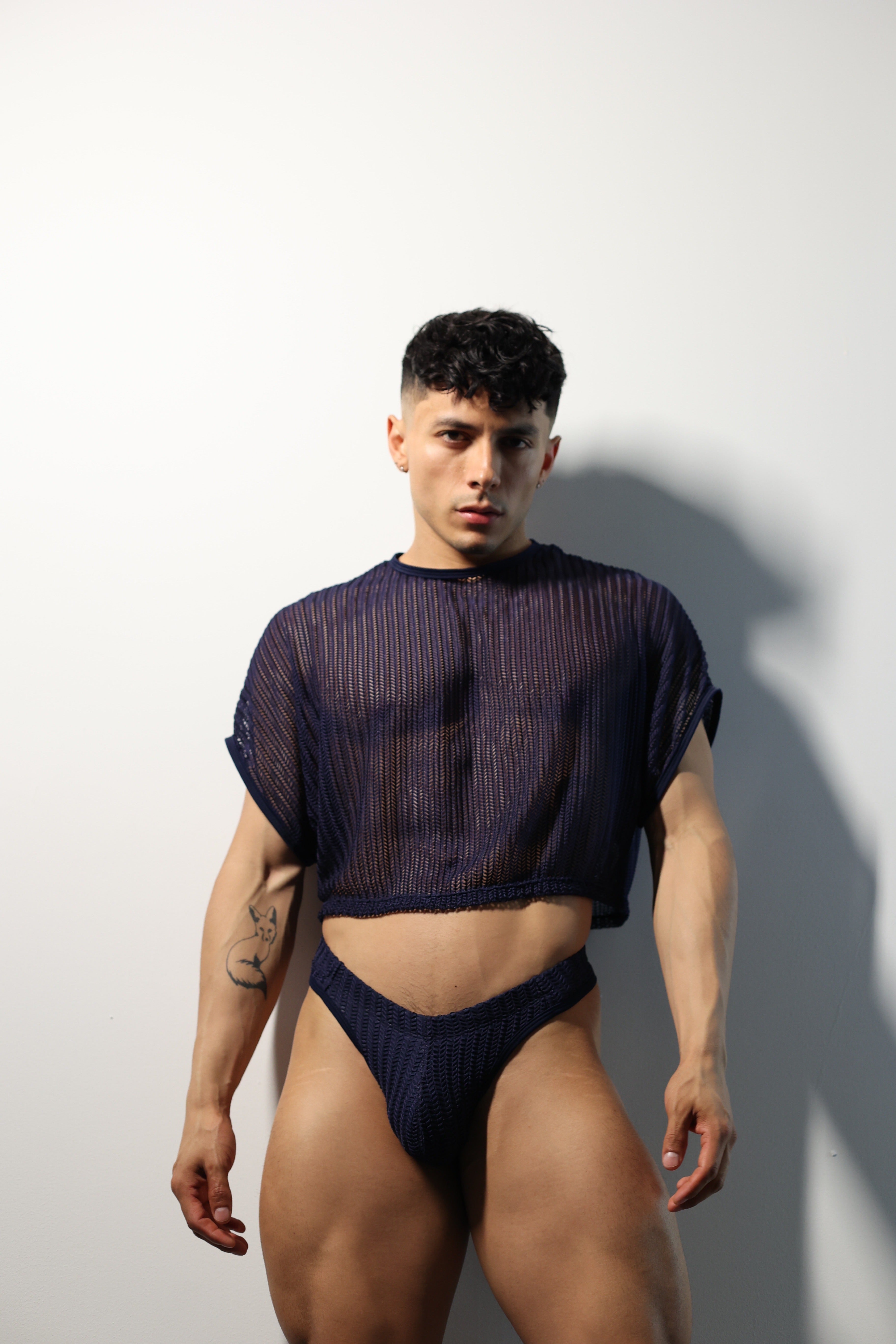 Fabian Divani modeling a sheer midnight blue mesh crop top and swim brief set—handmade men's swimwear—posed in front of a white brick wall