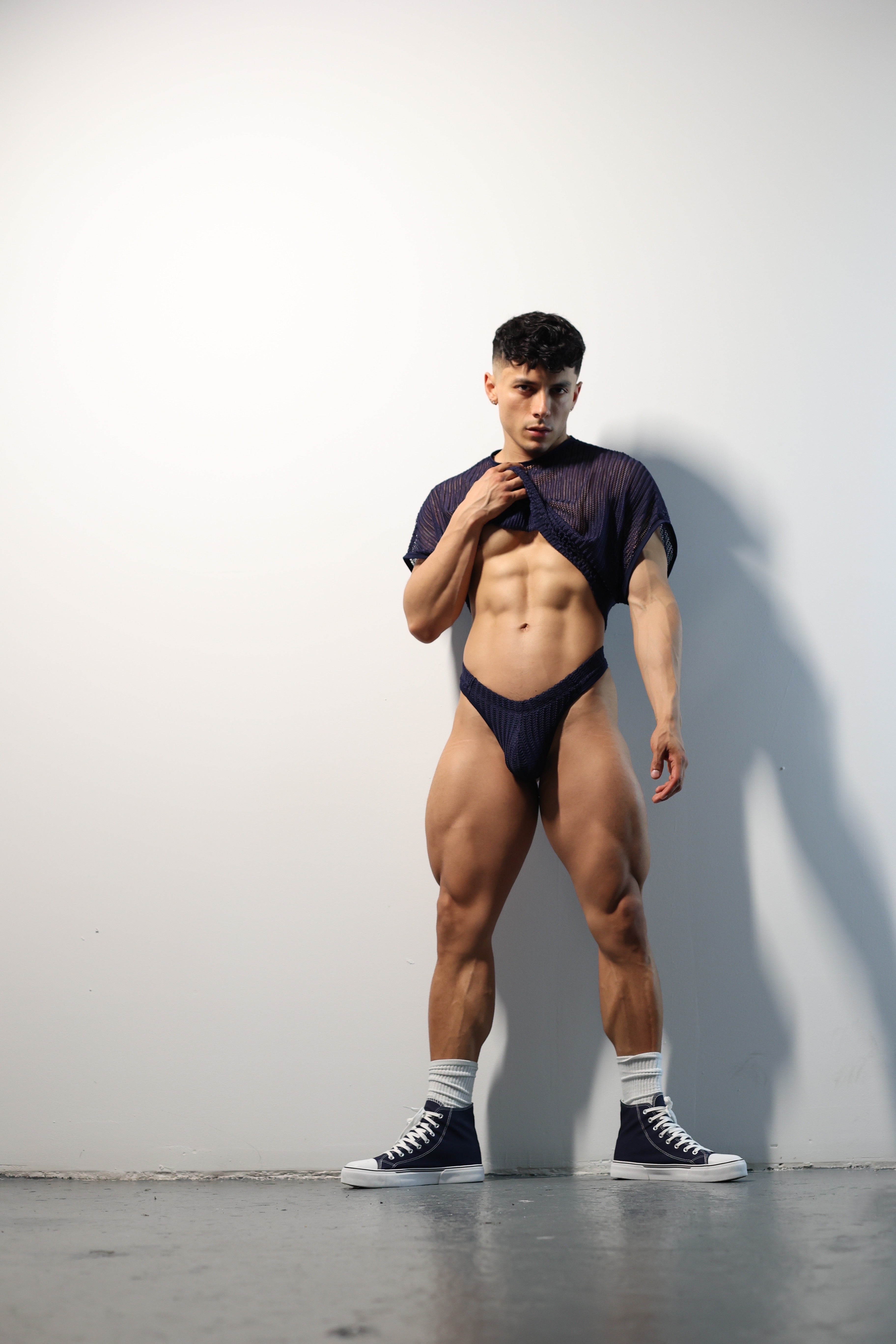 Fabian Divani modeling a sheer midnight blue mesh crop top and swim brief set—handmade men's swimwear—posed in front of a white brick wall
