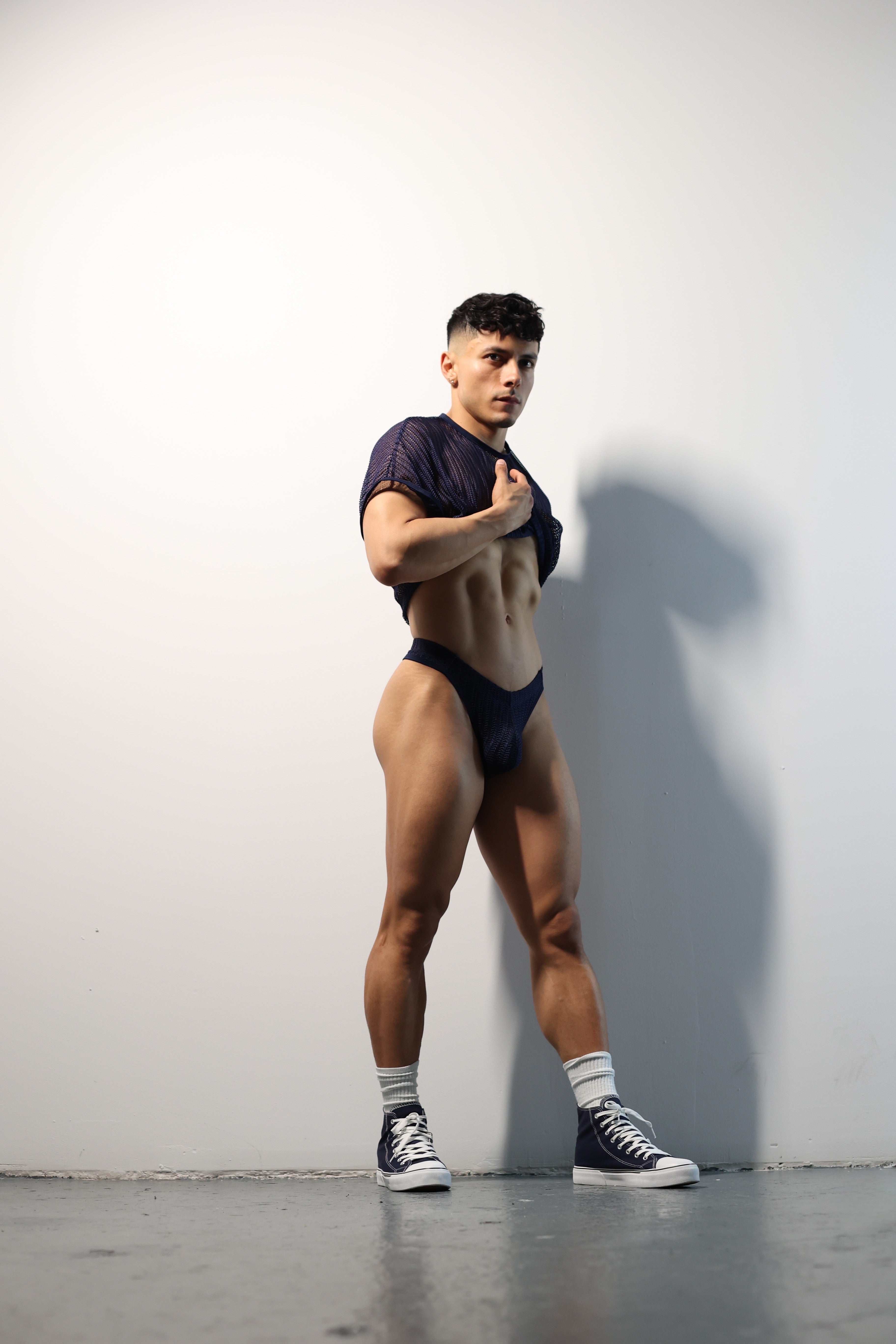 Fabian Divani modeling a sheer midnight blue mesh crop top and swim brief set—handmade men's swimwear—posed in front of a white brick wall