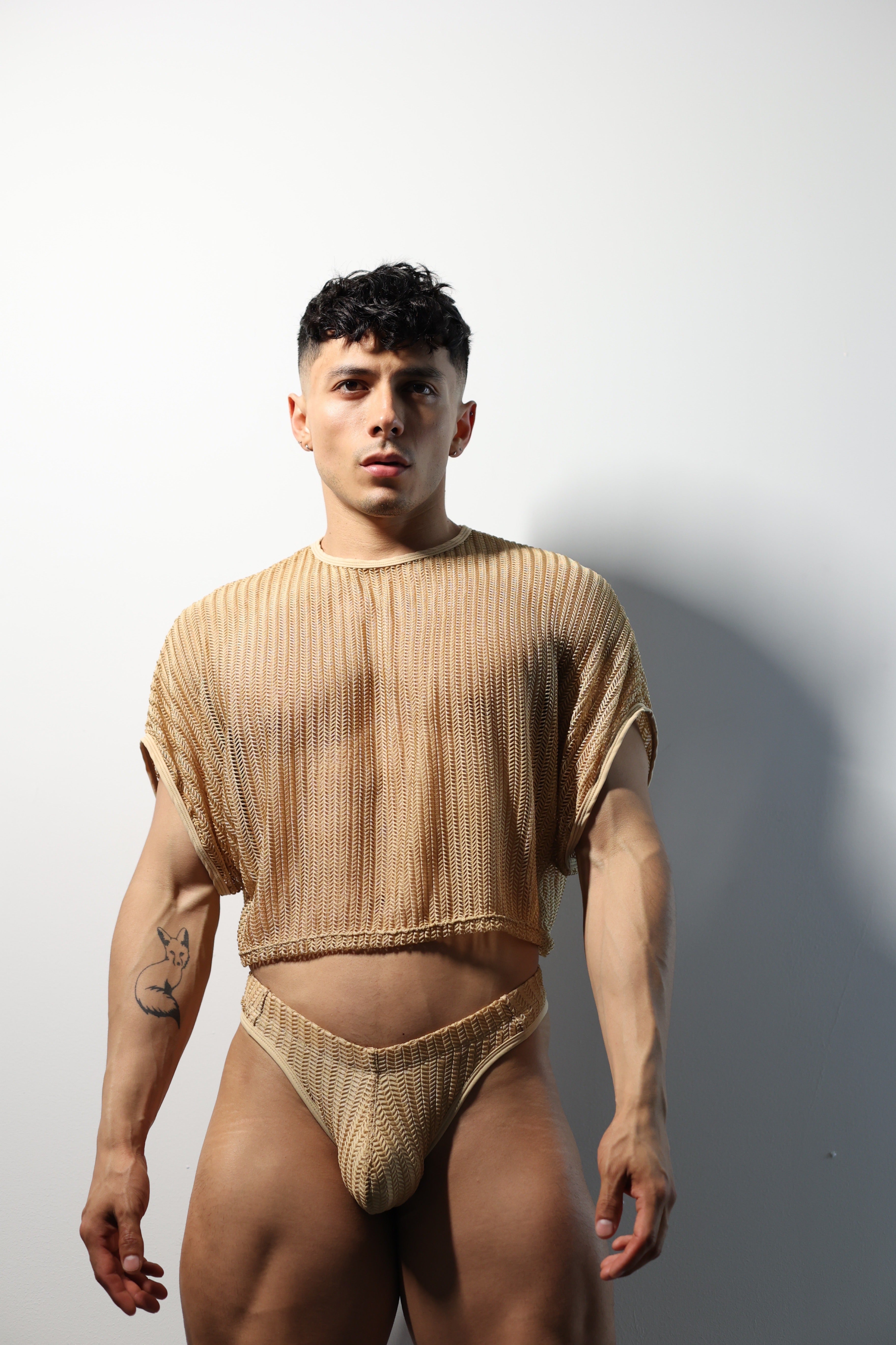 Fabian Divani modeling a netted natural crop top and swim brief set—handmade —posed in front of a white wall