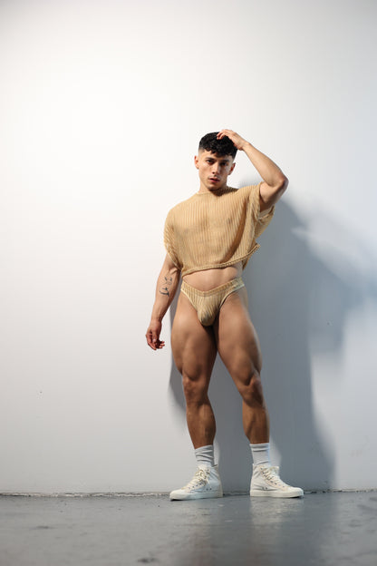 Fabian Divani modeling a netted natural crop top and swim brief set—handmade —posed in front of a white wall