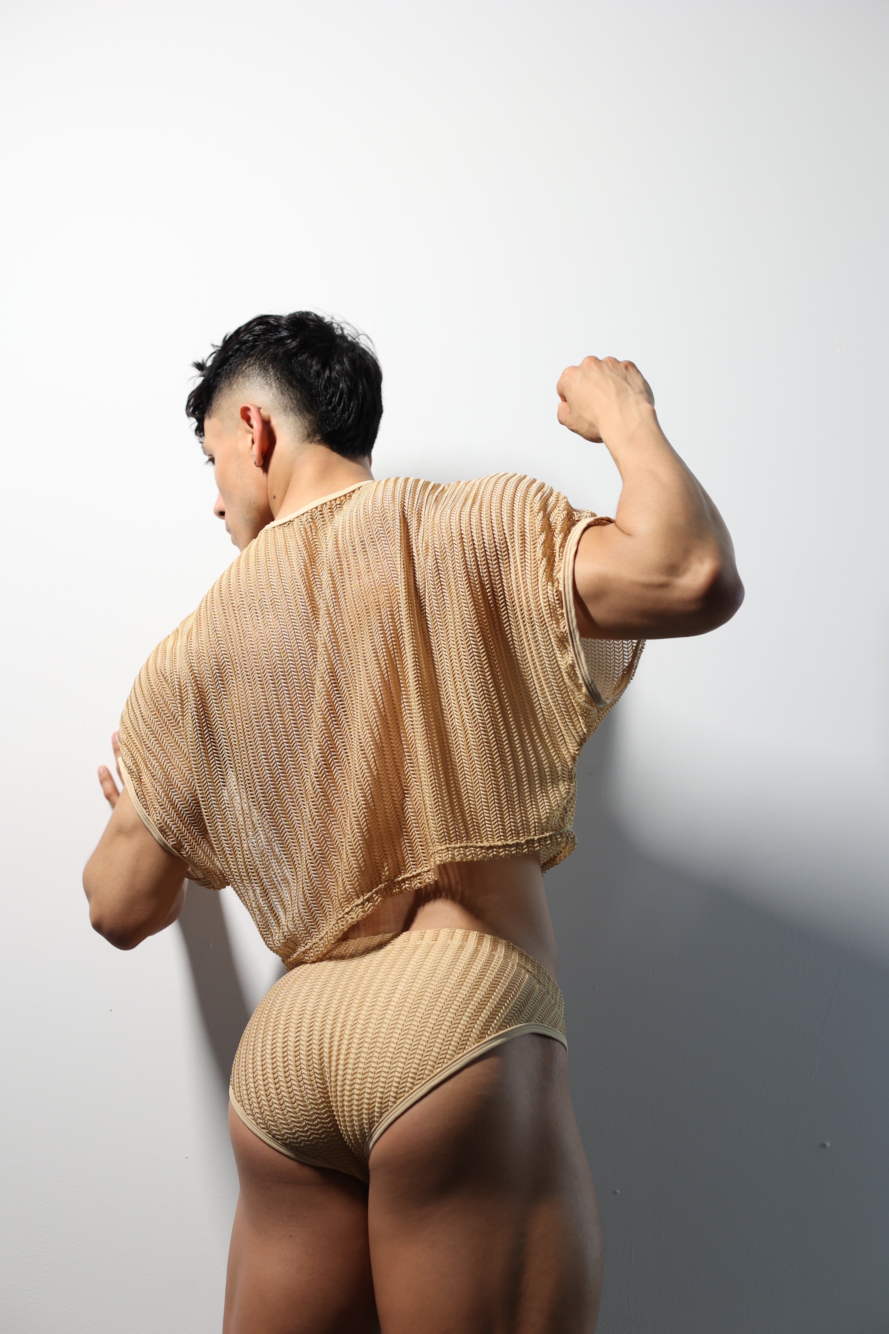 Fabian Divani modeling a sheer natural mesh crop top and swim brief set—handmade men's swimwear—posed in front of a white wall