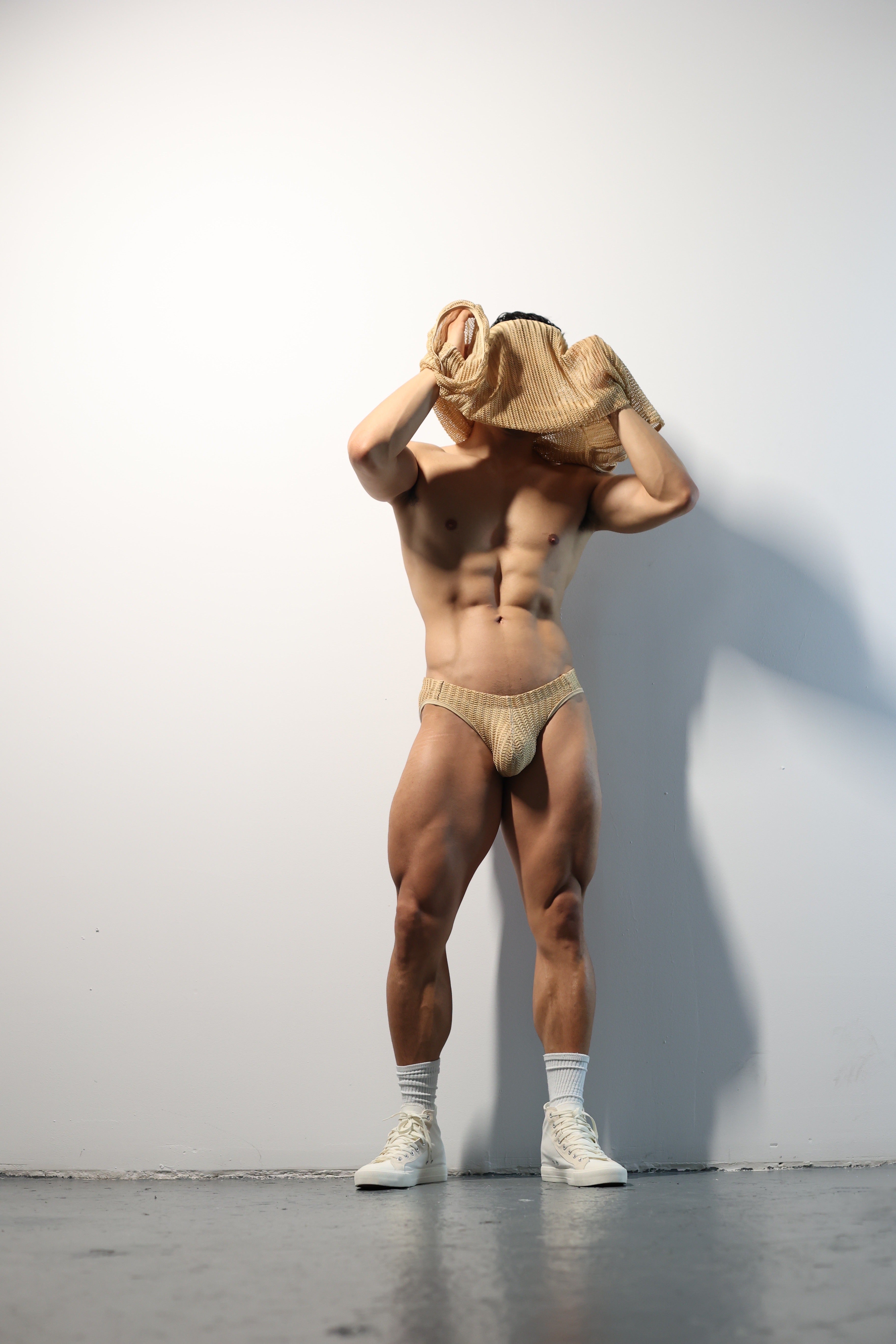 Fabian Divani modeling a sheer natural mesh crop top and swim brief set—handmade men's swimwear—posed in front of a white wall