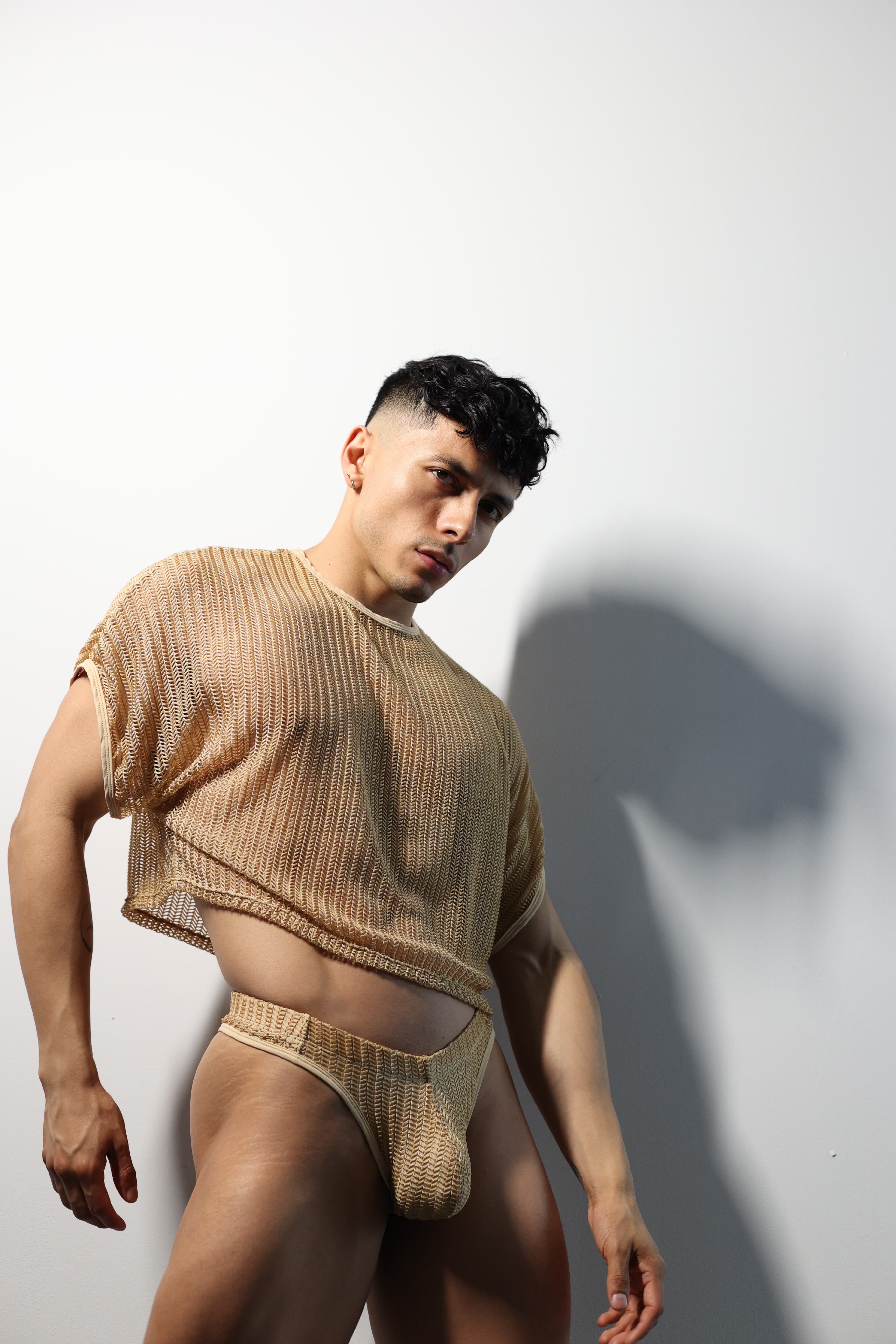 Fabian Divani modeling a sheer natural mesh crop top and swim brief set—handmade men's swimwear—posed in front of a white wall