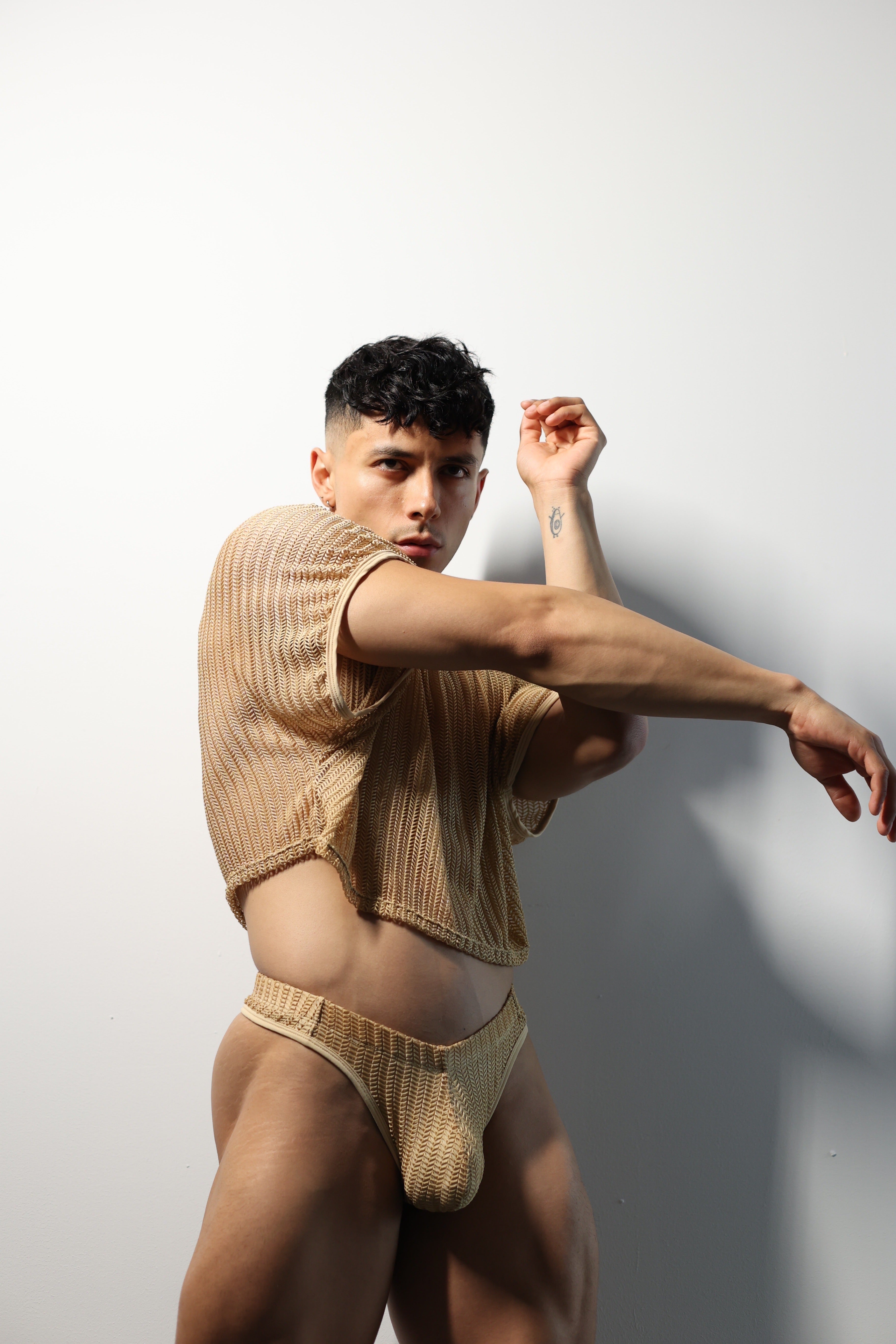 Fabian Divani modeling a sheer natural mesh crop top and swim brief set—handmade men's swimwear—posed in front of a white wall