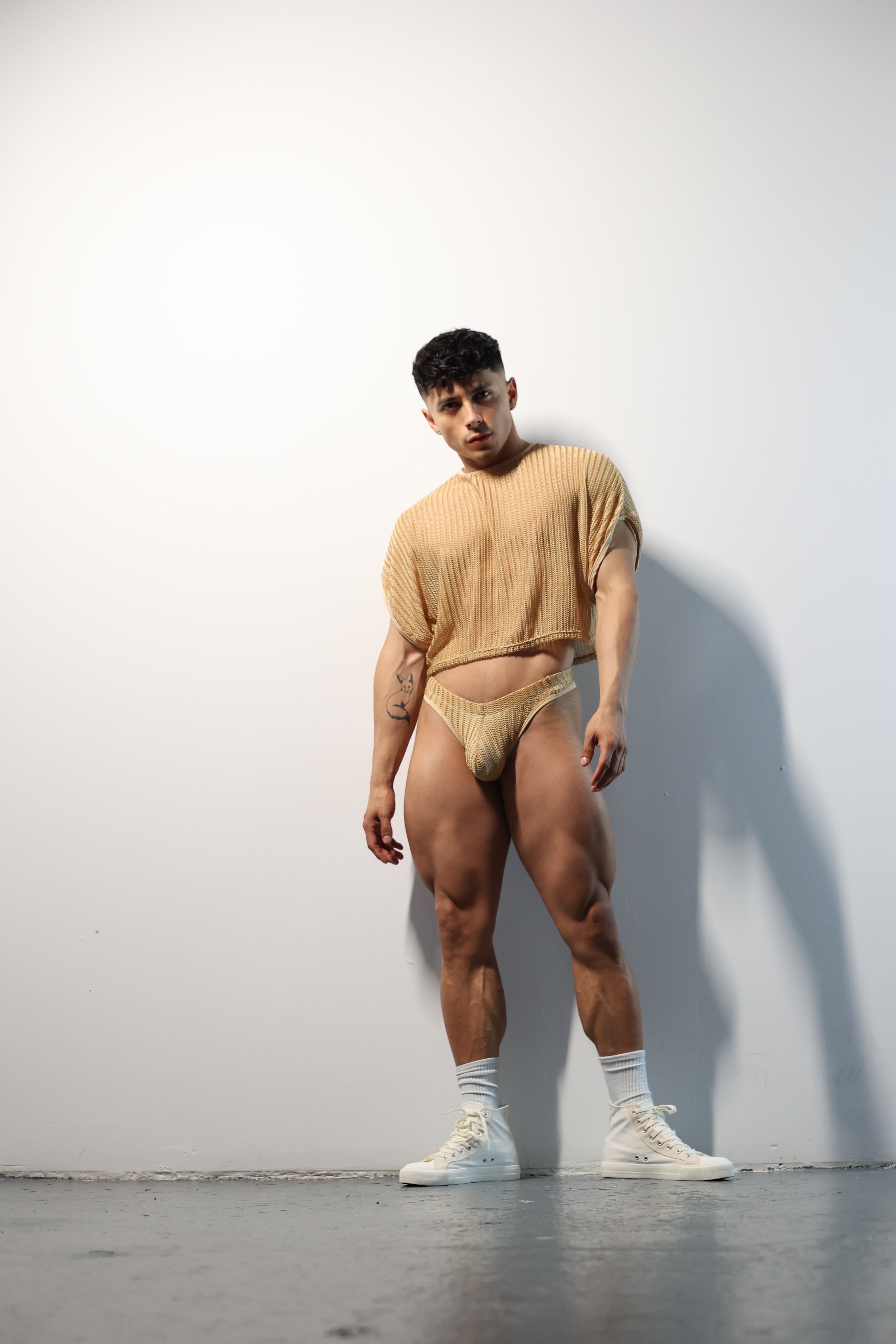 Fabian Divani modeling a sheer natural mesh crop top and swim brief set—handmade men's swimwear—posed in front of a white wall
