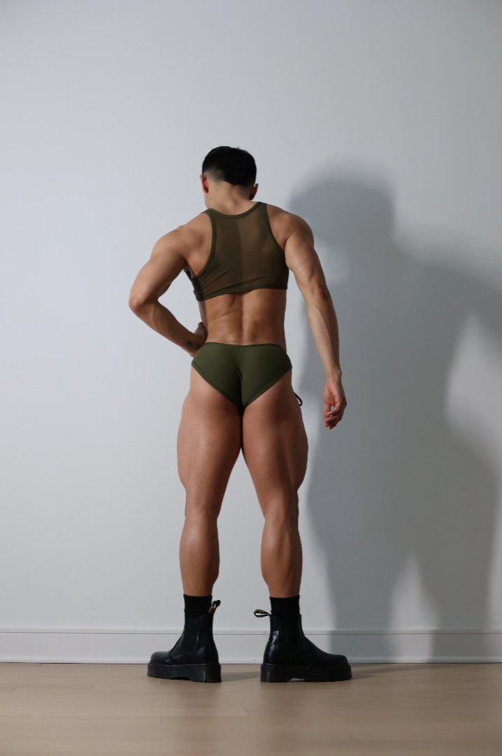 Fabian Divani modeling a sheer olive crop top and swim brief set—handmade men's swimwear—posed in front of a white wall