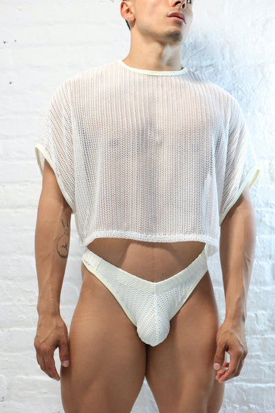 Fabian Divani modeling a sheer white mesh crop top and swim brief set—handmade —posed in front of a white brick wall