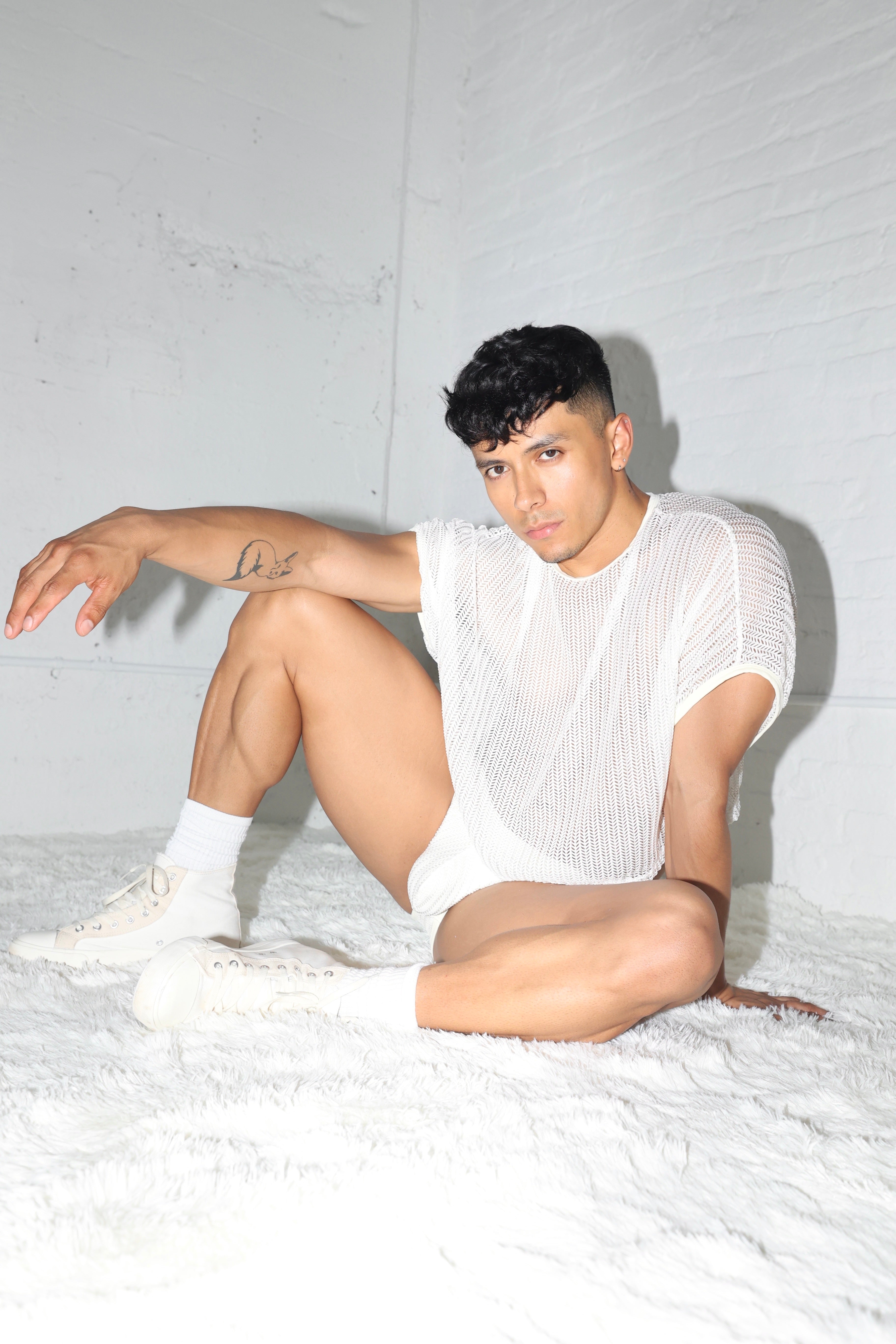 Fabian Divani modeling a sheer white mesh crop top and swim brief set—handmade by Fabi —posed in front of a white brick wall