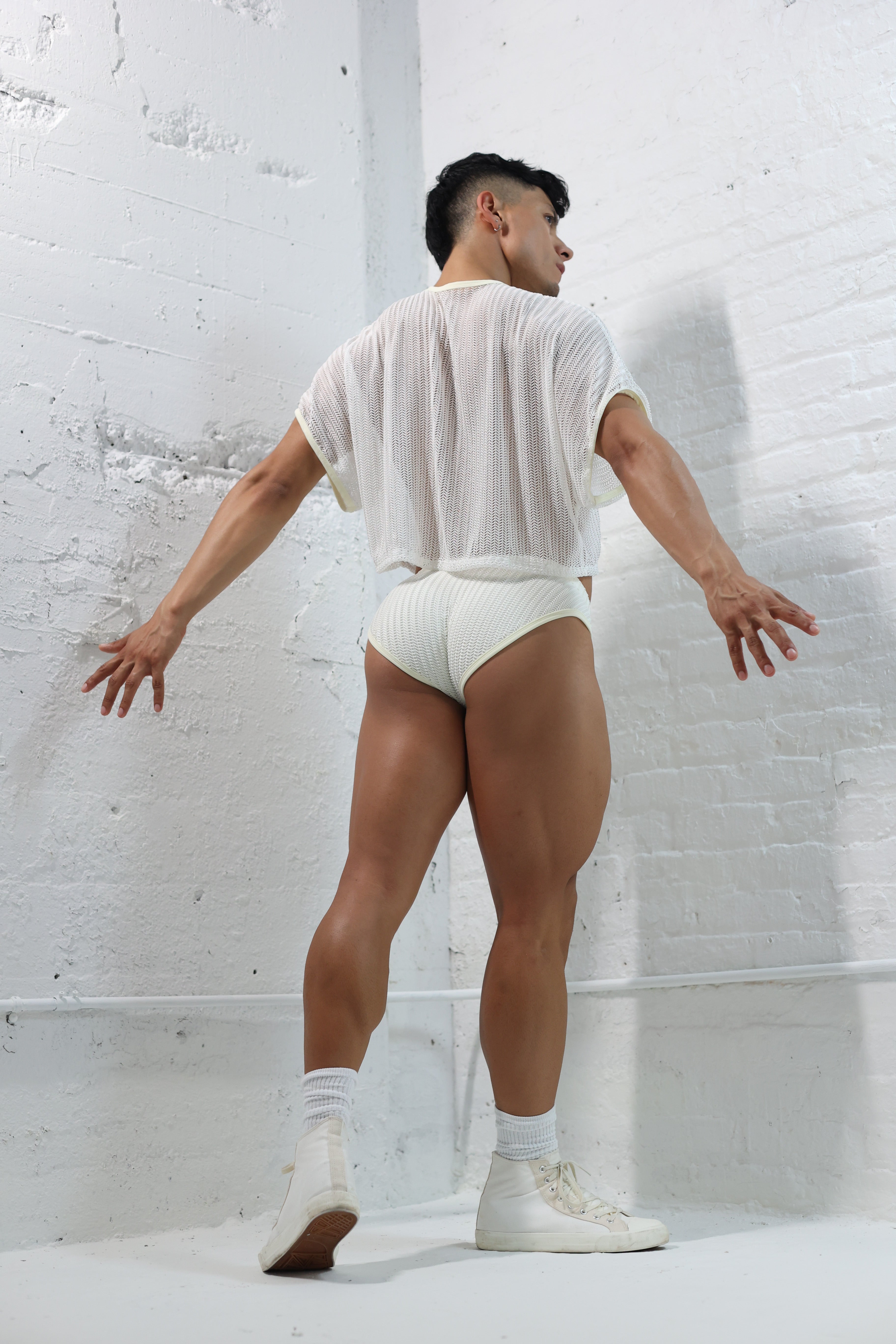 Fabian Divani modeling a sheer white mesh crop top and swim brief set—handmade men's swimwear—posed in front of a white brick wall